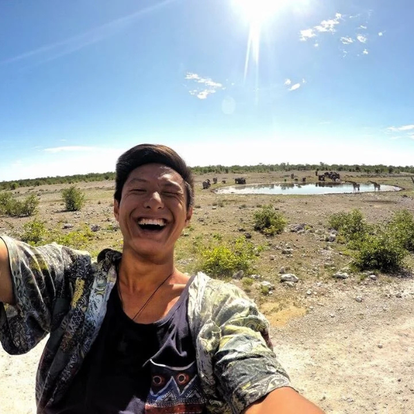 Person taking a selfie in a savannah landscape with a watering hole and grazing animals in the distance under a bright blue sky with a few clouds.