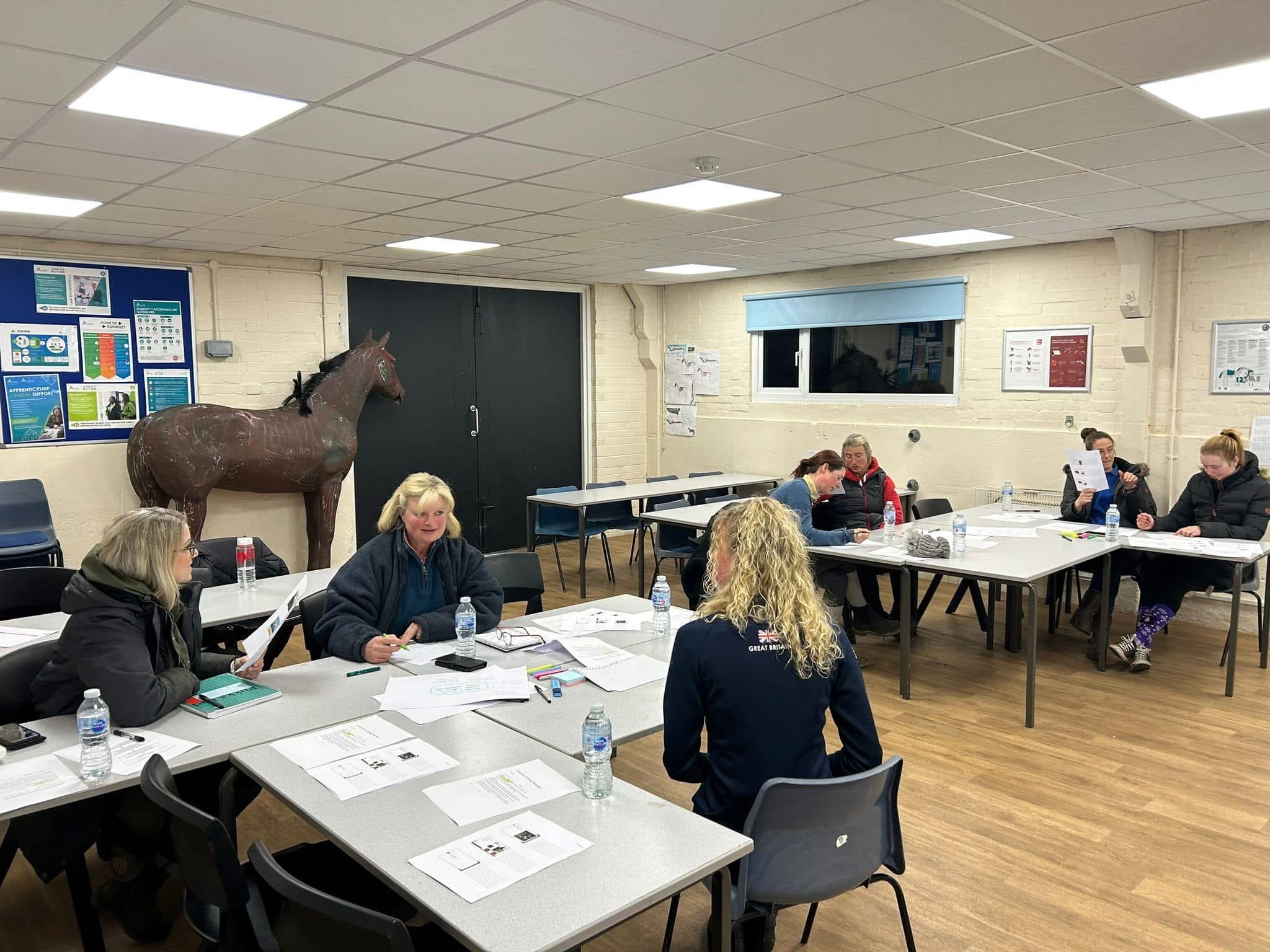 People sitting at tables in a meeting room, with documents and water bottles, and a horse mannequin in the background.
