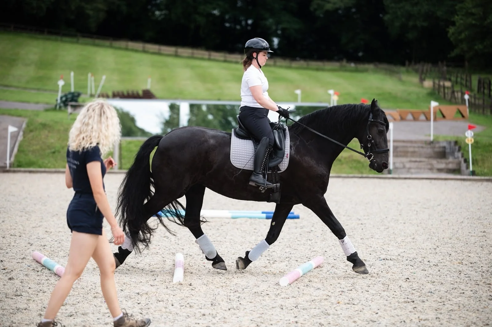 A woman riding a black horse through a riding course, guided by a woman with curly blond hair.