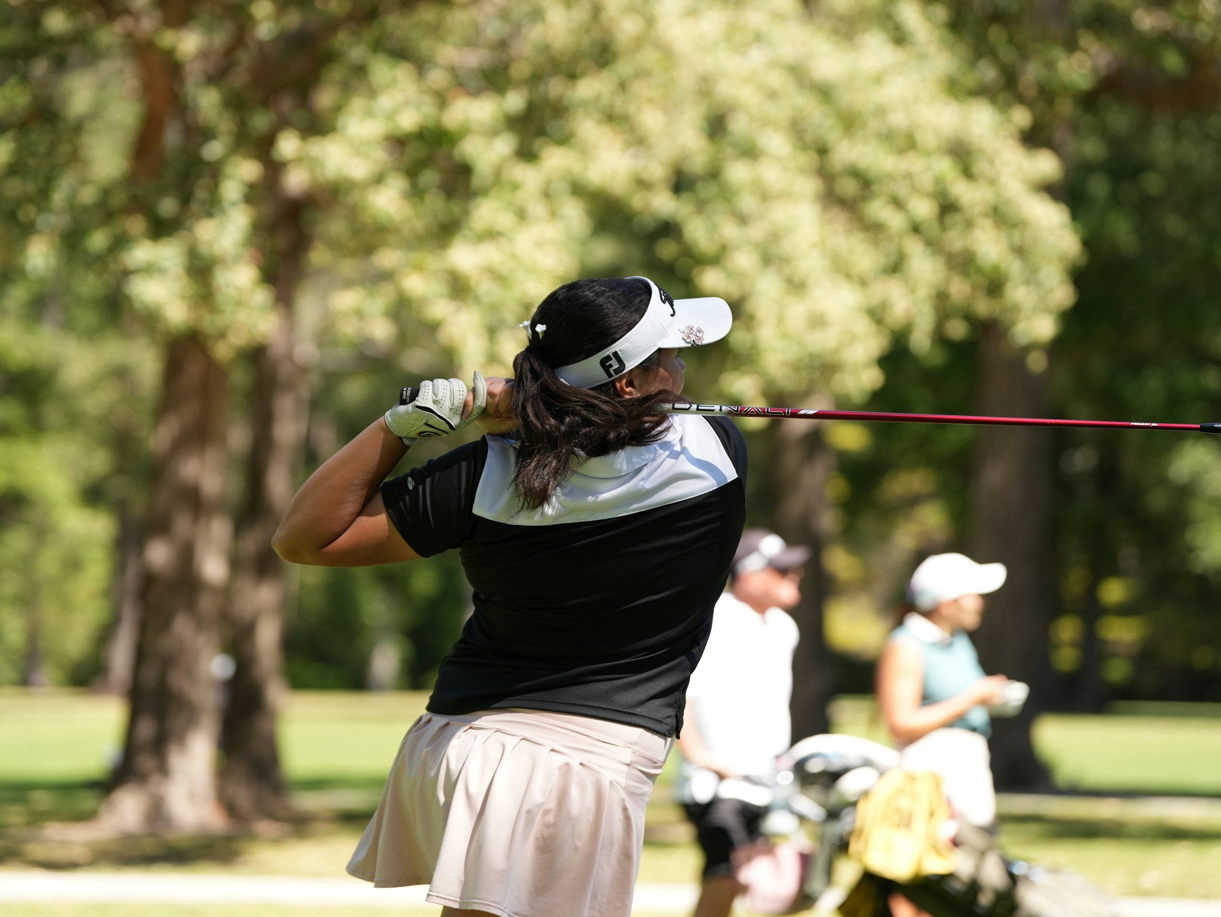 A woman in golf attire, including a visor, is swinging a golf club in a park with trees in the background. She is wearing a black and white shirt and beige shorts. Other people and golf carts are visible in the background.