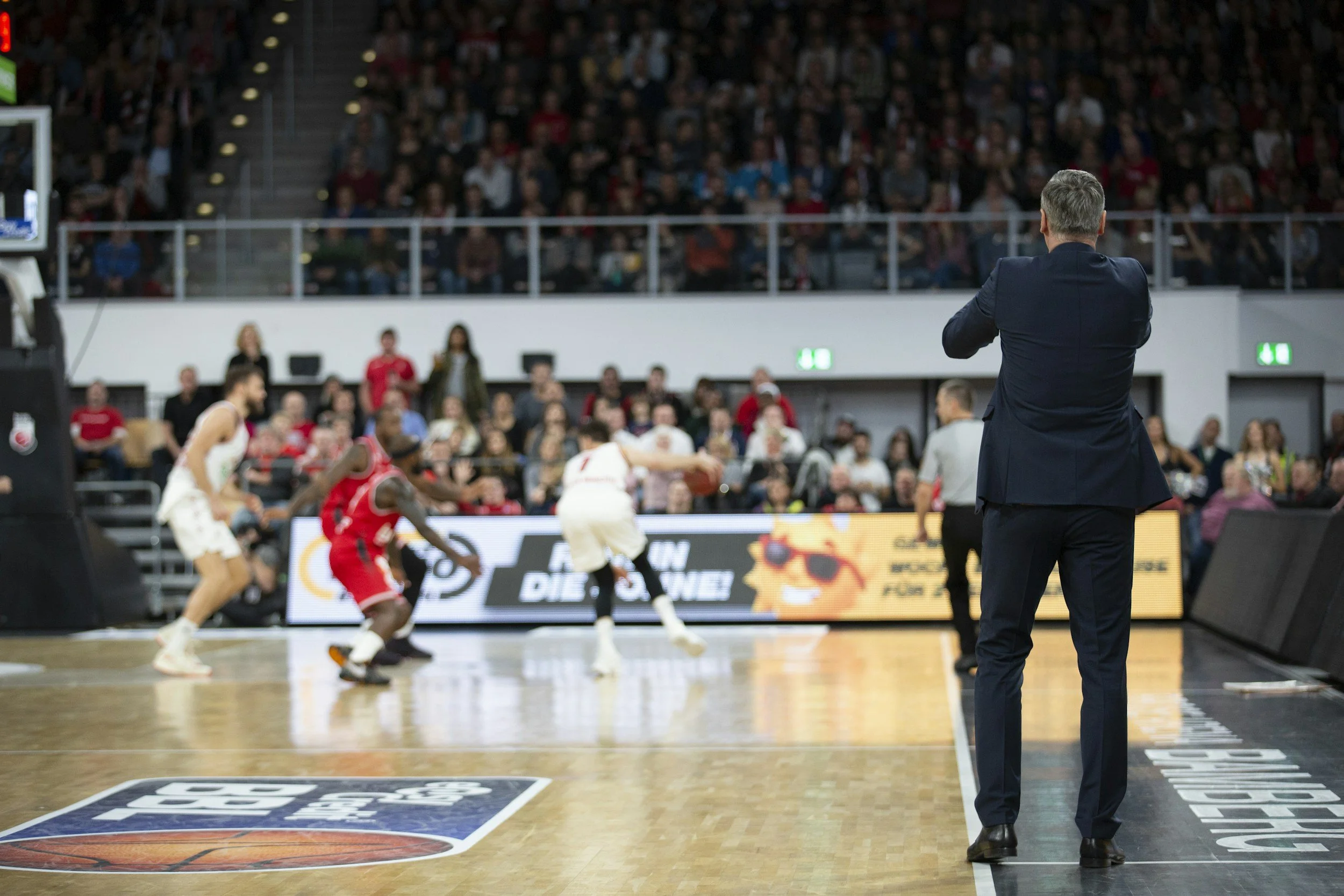 A basketball coach with gray hair in a dark suit stands with arms crossed, watching a game in a large indoor arena filled with spectators, some players visible on the court.
