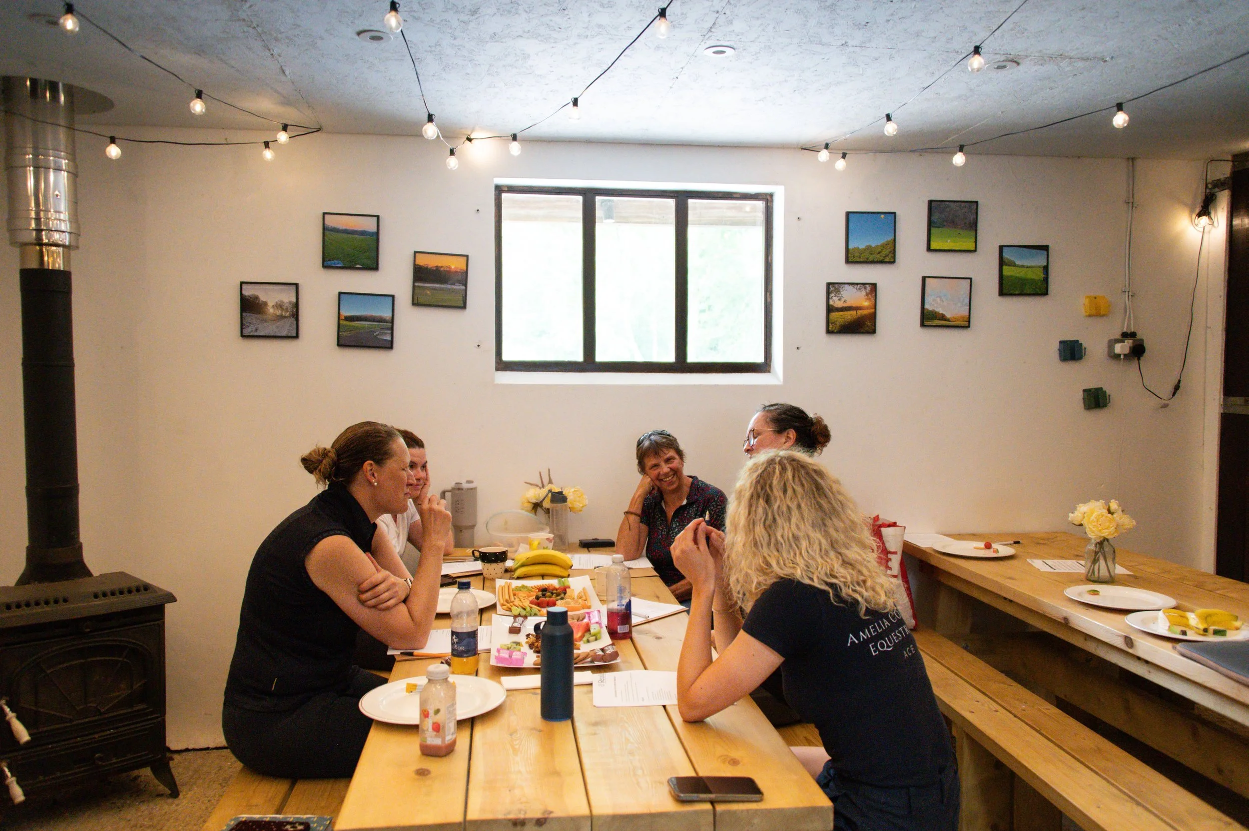 People sitting around a table sharing a meal and laughing in a cozy indoor space. The room has string lights on the ceiling, framed pictures of landscapes on the walls, and a window letting in natural light.