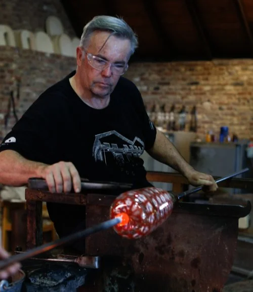 Man wearing glasses and a black T-shirt glassblowing a red and white glass object in a workshop.