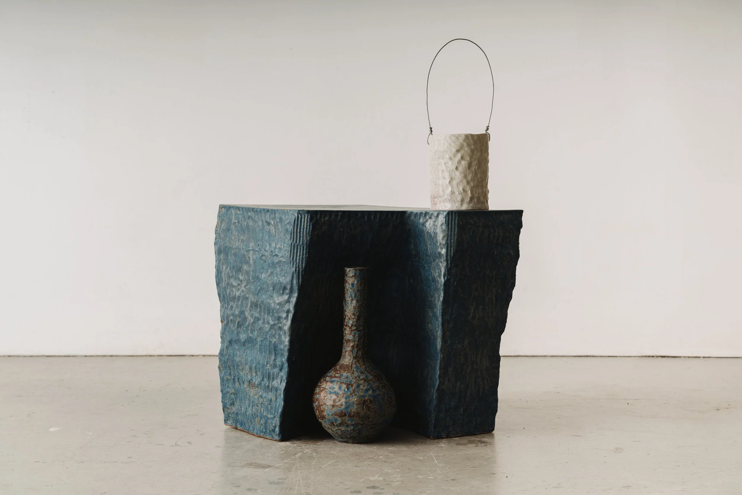 Minimalist sculpture composed of irregularly shaped, rough-textured blocks in shades of blue, beige, and brown, stacked in a vertical arrangement with a wire loop at the top.