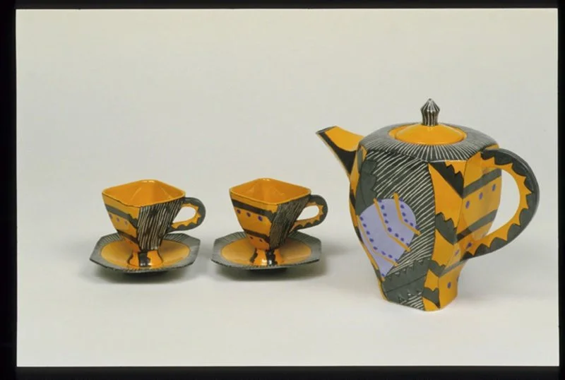 A ceramic tea set with one teapot and two teacups with saucers, decorated in abstract patterns of black, yellow, orange, and purple, displayed against a white background.
