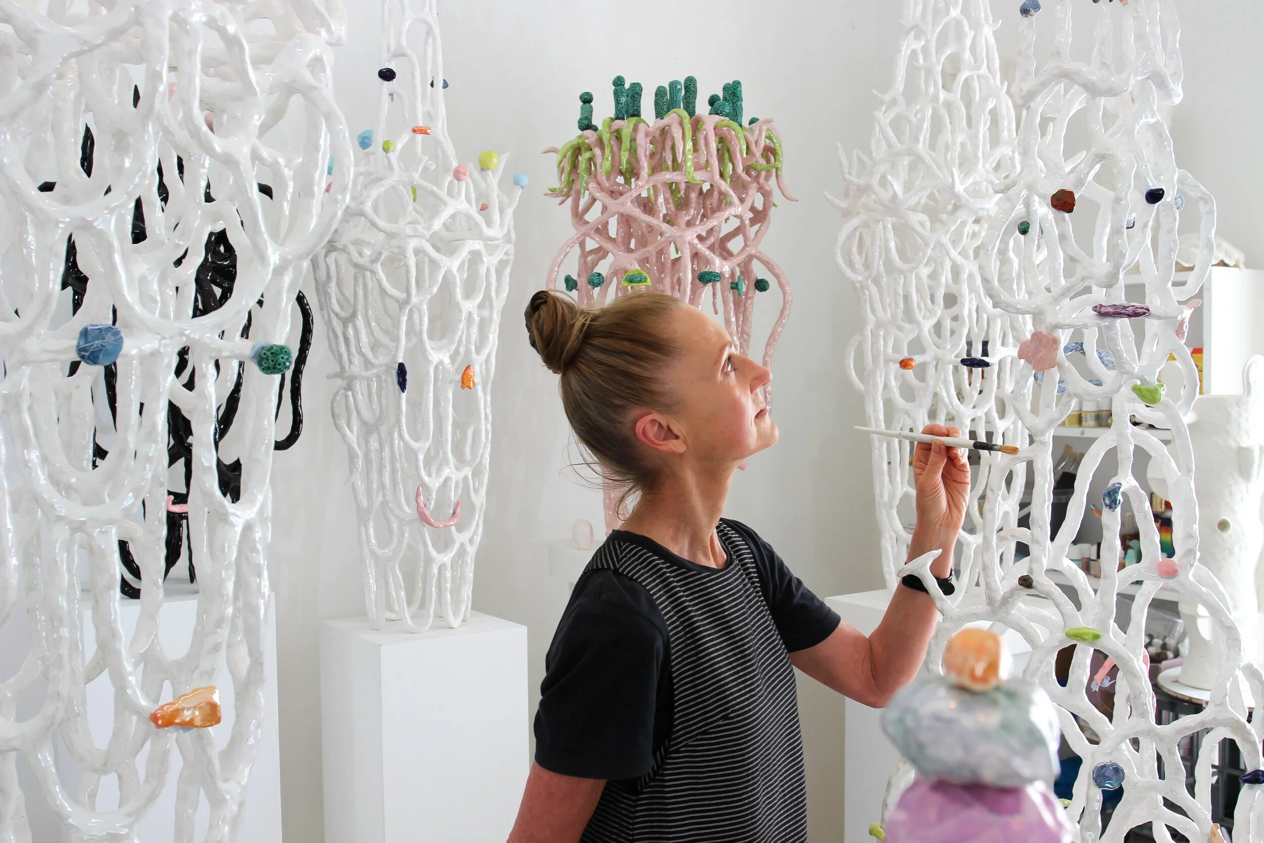 A woman in a striped shirt painting abstract, looped, white sculptures with colorful accents in an art studio.