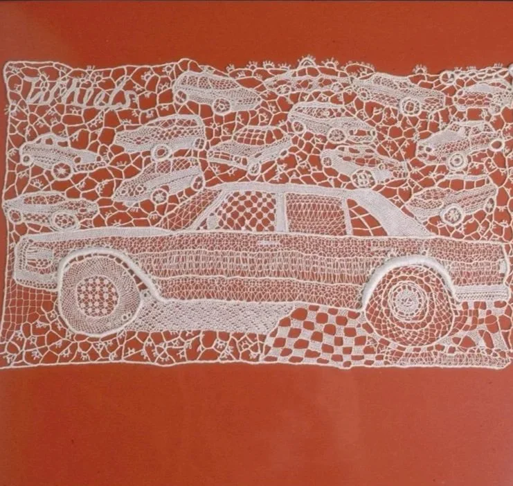 White lace artwork depicting a vintage car with cars above it, set against a reddish-orange background.