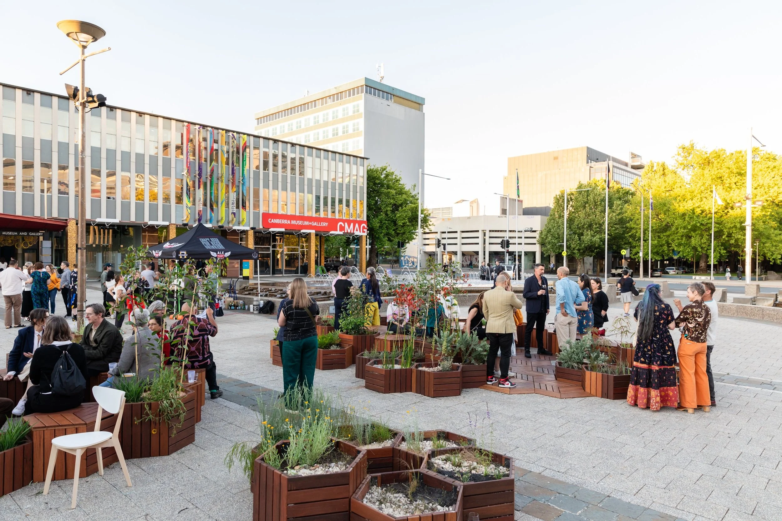 People gathered in an outdoor plaza with plants in wooden planters, in front of the Canberra Museum and Gallery, with tall buildings and trees in the background.