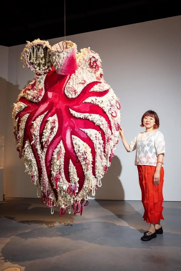 A woman standing next to an art installation resembling a human heart made of various flowers and decorative elements, displayed indoors.