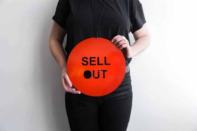 Person holding a red circular sign that reads 'SELL OUT' against a plain white wall.