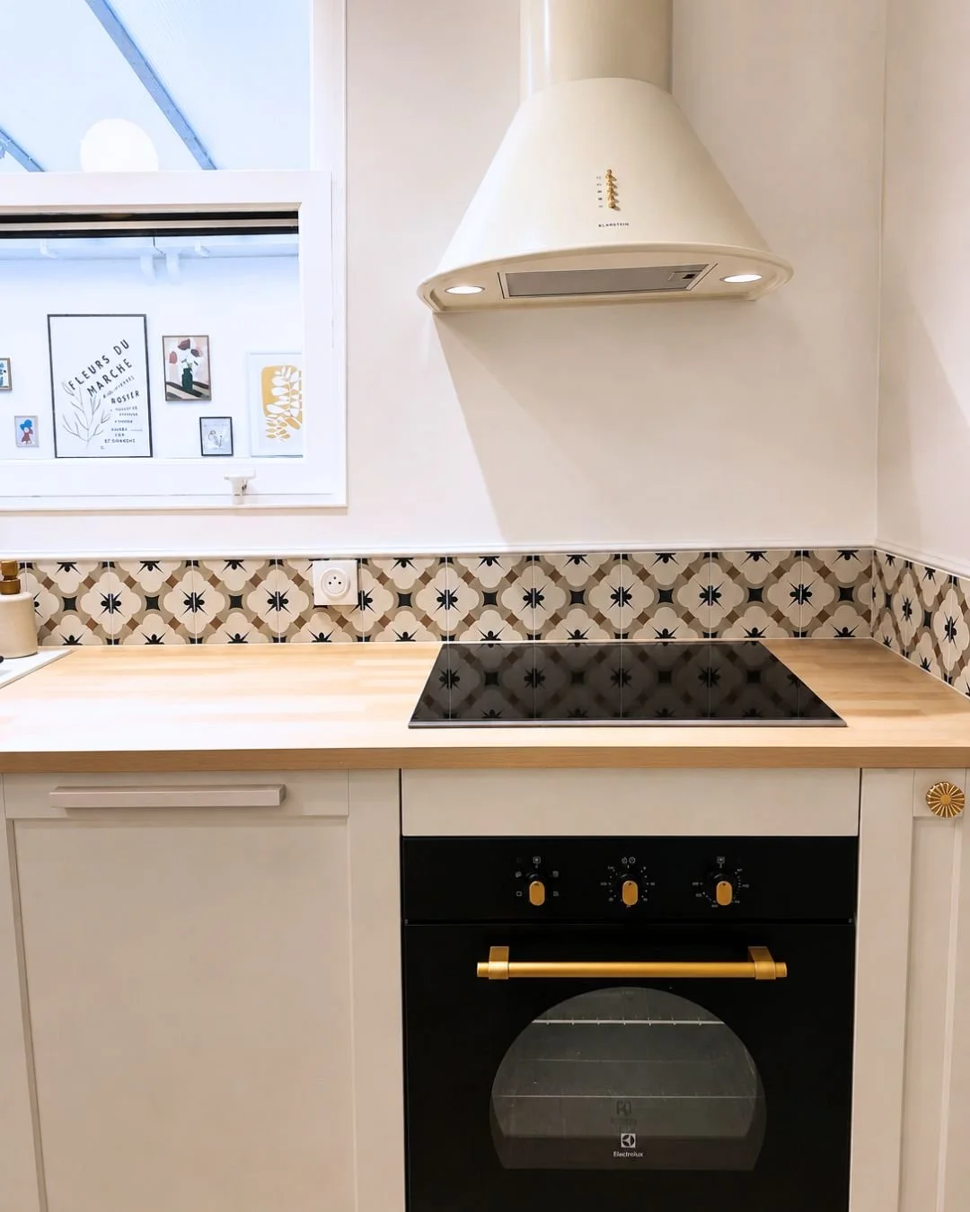Cuisine avec four électrique noir, plaques de cuisson, crédence en carreaux décoratifs, hotte blanche, fenêtre avec déco murale.