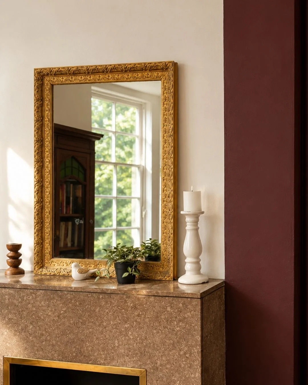 Miroir encadré doré au-dessus d'une cheminée en marbre, décorée de plantes, d'une bougie blanche sur un chandelier en céramique, et d'une petite sculpture en forme d'oiseau.