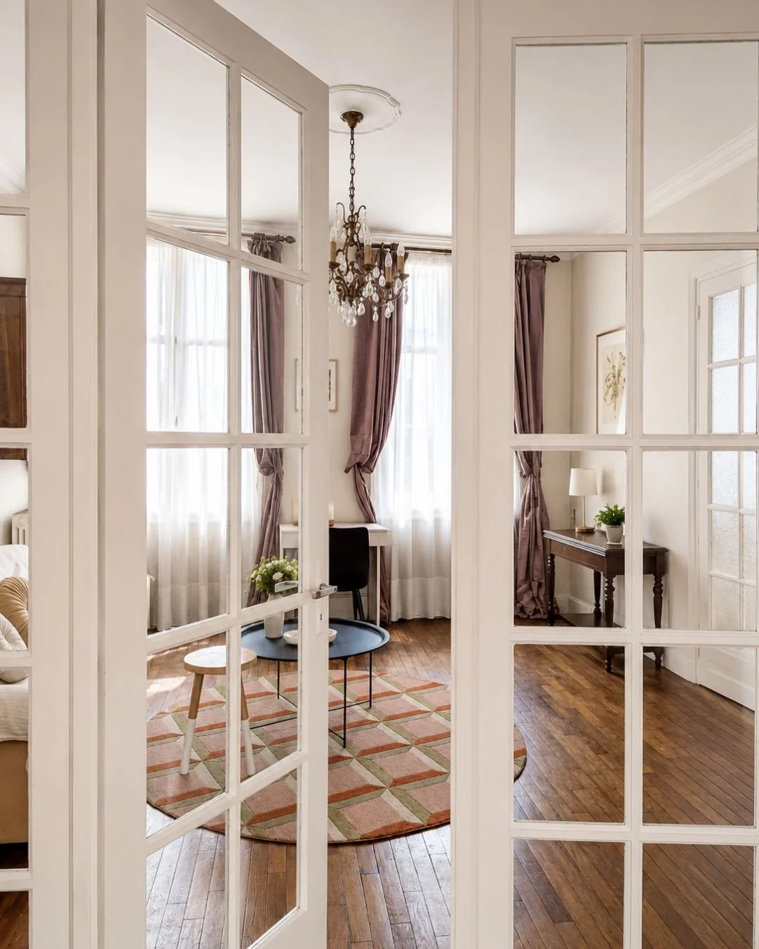 Intérieur d'une pièce vue à travers une porte en verre avec plusieurs panneaux. La pièce dispose de murs blancs, de rideaux violets, d'un chandelier, d'une table ronde noire, d'une petite table en bois, d'une lampe, et de rideaux derrière de grandes fenêtres.