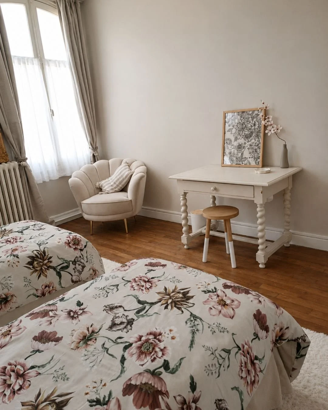Chambre l'Envol avec deux lits à fleurs, un fauteuil beige près de la fenêtre, un bureau blanc avec un petit tabouret en bois, une œuvre d'art et un vase avec des fleurs roses.