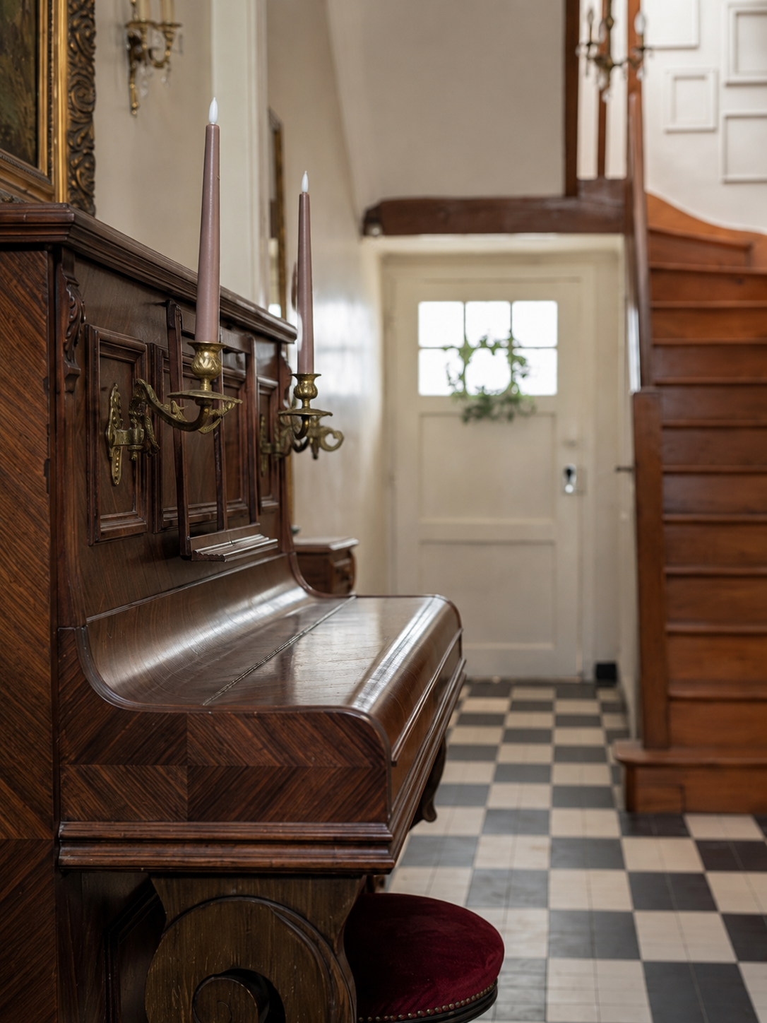 Entrée Maison Tourterelle. Un piano en bois avec des chandeliers accrochés dessus dans une entrée ou un couloir, avec une porte blanche en fond et un escalier en bois à droite.