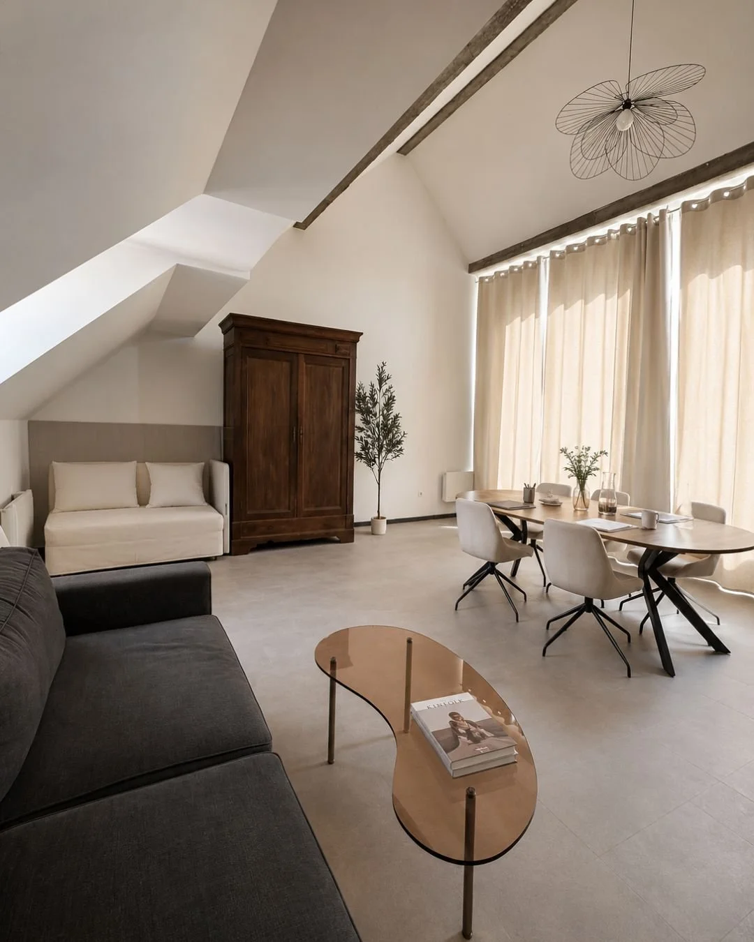 Salon moderne dans le loft Le Perchoir avec canapé gris, table en bois, rideaux beiges, armoire en bois foncé, plantes vertes, plafonnier en forme de papillon en métal, grand espace lumineux.