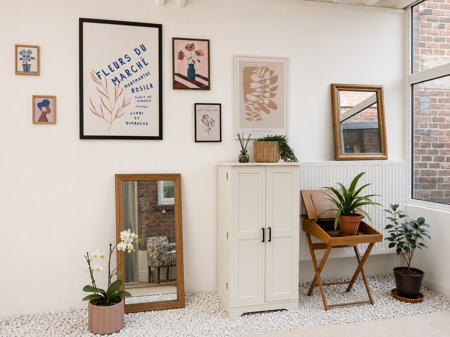 Intérieur de la véranda avec des murs blancs, décorations florales en cadres, miroir, plantes en pot, meuble blanc et panneau radiateur.