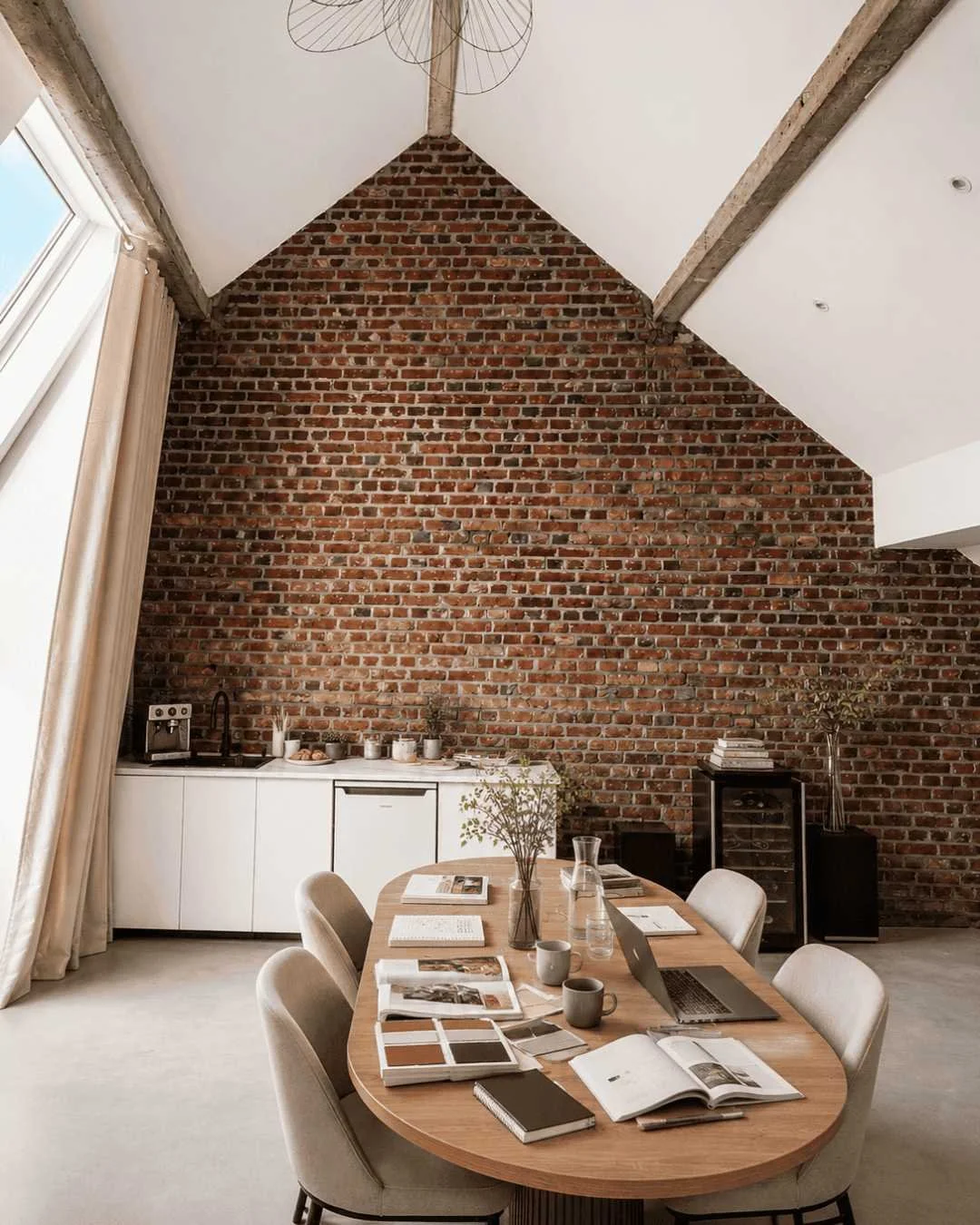 Salle à manger loft Perchoir avec une grande table en bois, six chaises beige, une cheminée en briques apparentes, un petit bar avec une vitrine, des livres, des plantes en décoration, et des velux avec rideaux lumineux.