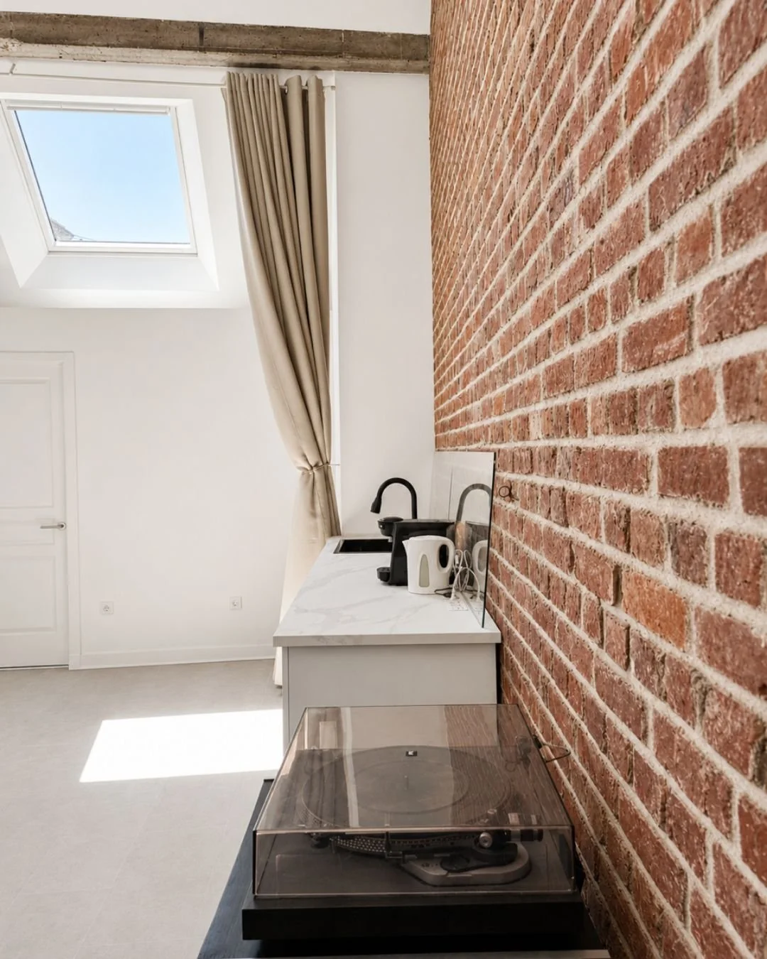 Coin cuisine moderne loft Le Perchoir avec évier noir, appareils électroménagers blancs, mur en briques rouges, fenêtre de toit avec rideau beige, platine vinyle.
