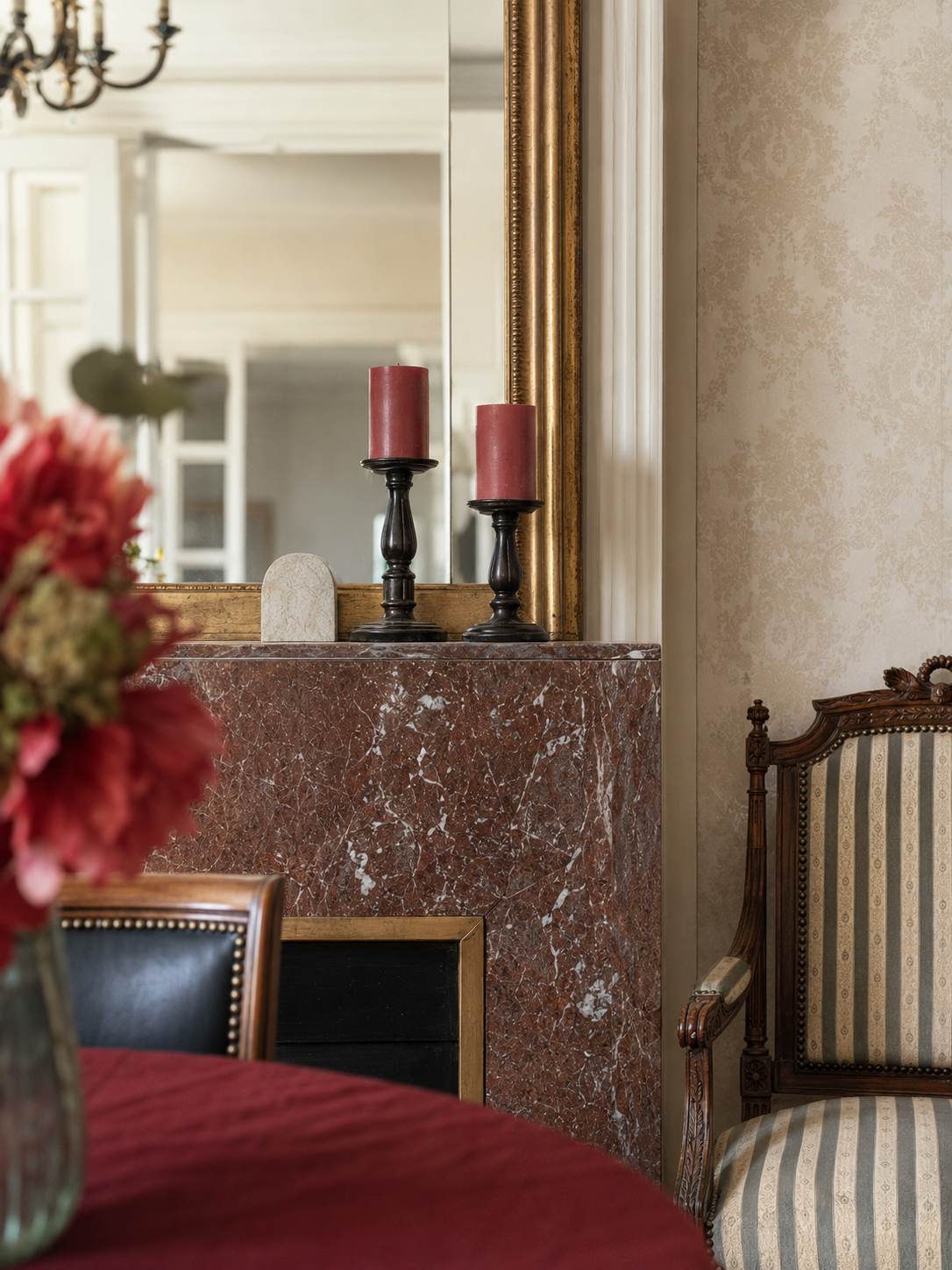Salle à manger avec une cheminée en marbre rouge, un grand miroir avec un cadre doré, deux bougies rouges sur des chandeliers noirs, une chaise en bois avec un tissu rayé beige et une table recouverte d'une nappe rouge, décor classique élégant.