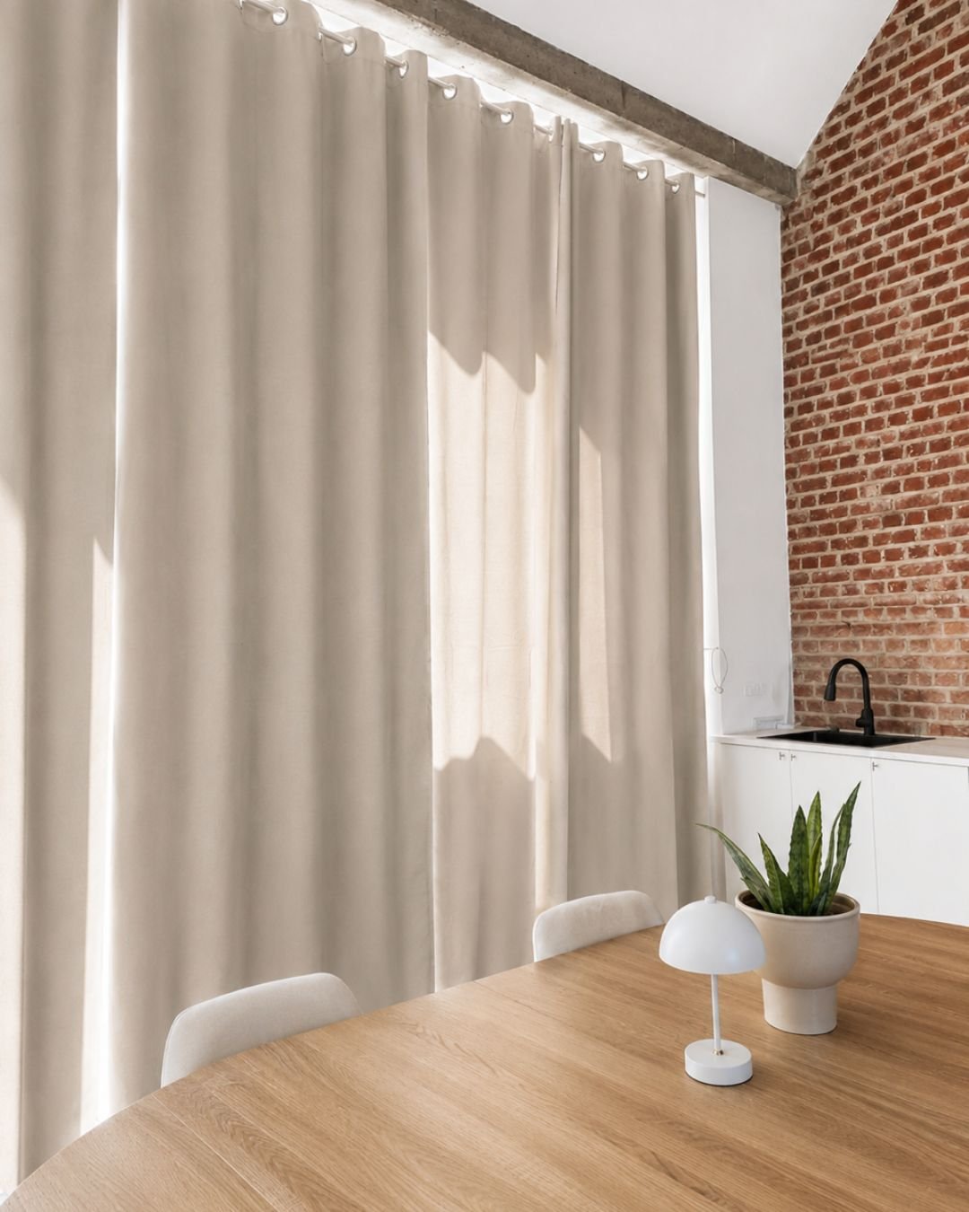 Une table en bois avec une plante verte en pot, une petite lampe blanche et des chaises entourant la table. À l'arrière, un mur en briques rouges, un comptoir avec un évier et une robinetterie noire, et de longues rideaux beige couvrant une fenêtre o