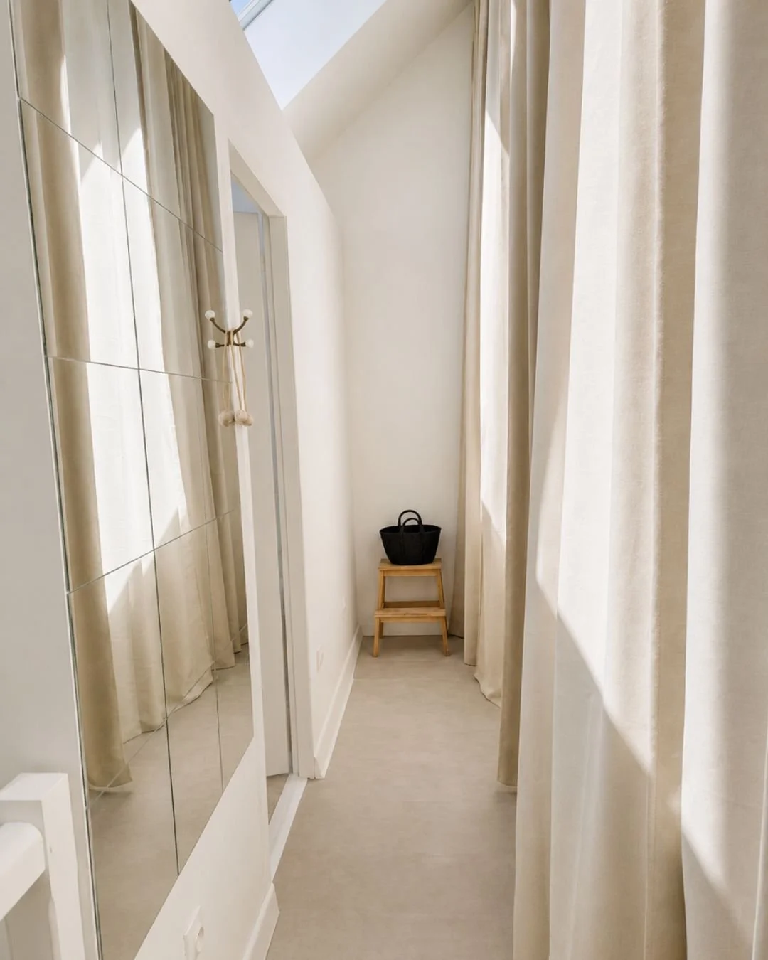 Entrée loft Le Perchoir avec murs blancs, rideaux crème, un miroir partiel, un porte-manteau, un petit escabeau en bois avec un sac noir dessus, et un plafond incliné avec une fenêtre ou puits de lumière