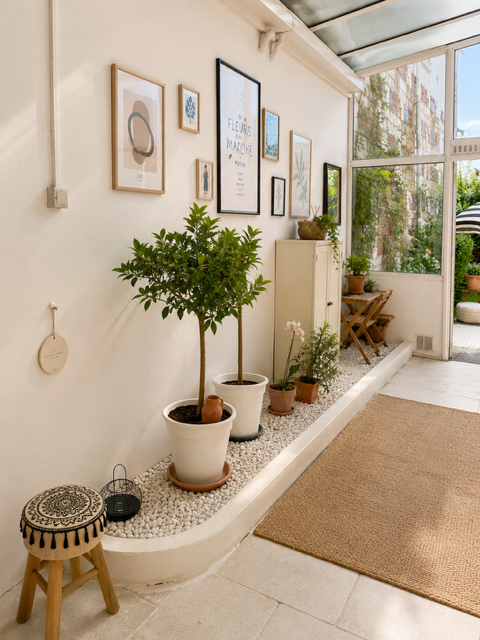 Intérieur véranda lumineux avec une jardinière contenant plusieurs plantes, œuvres d'art encadrées sur un mur, une petite table et une porte-fenêtre donnant sur un jardin avec des plantes