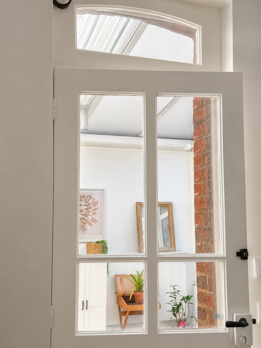 Vue à travers la porte de la cuisine en verre divisée en plusieurs panneaux, montrant un intérieur lumineux avec des décorations végétales, tableaux et une colonne en briques apparentes.
