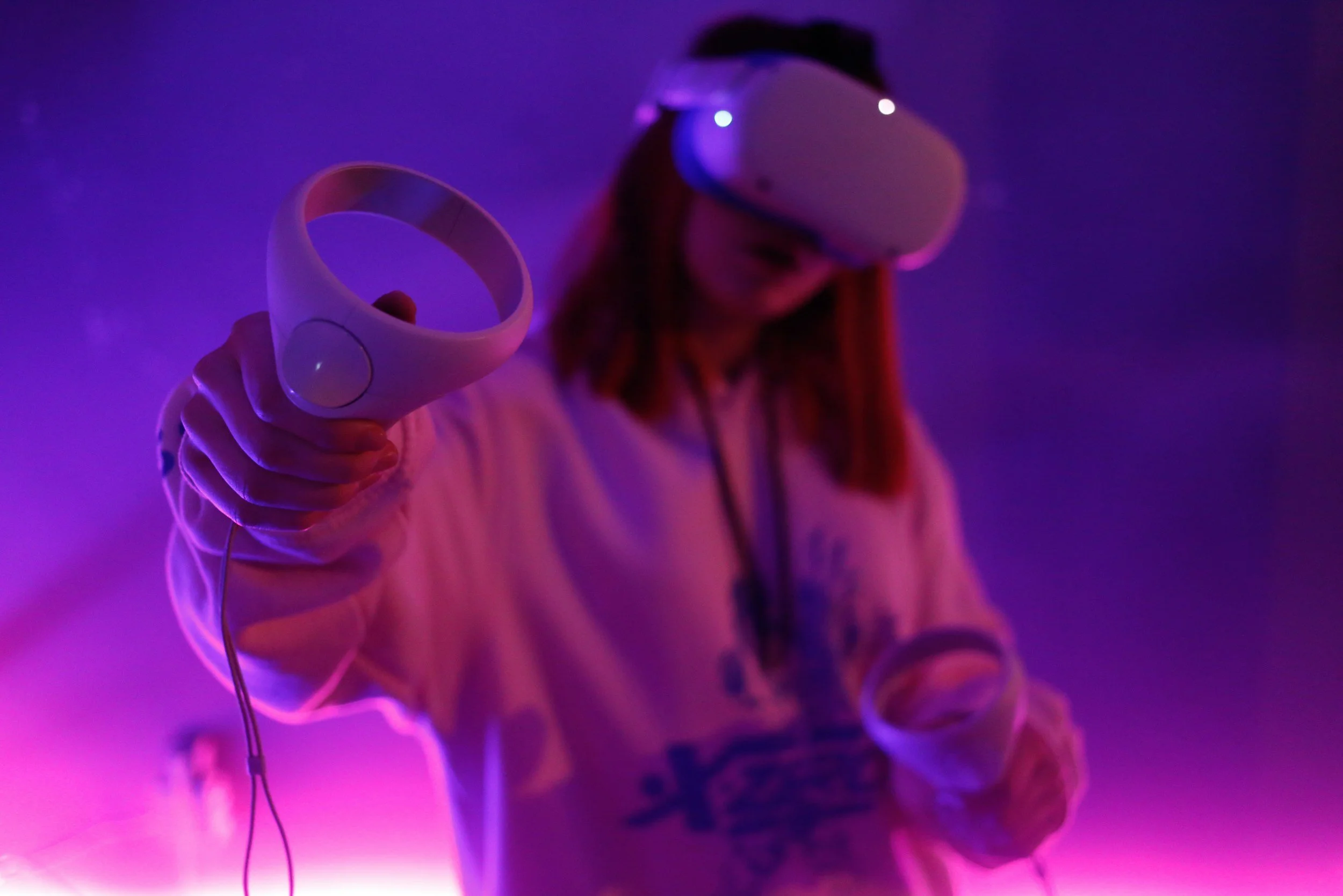 A person wearing a virtual reality headset holding a wire controller in a dimly lit space with purple and pink lighting.