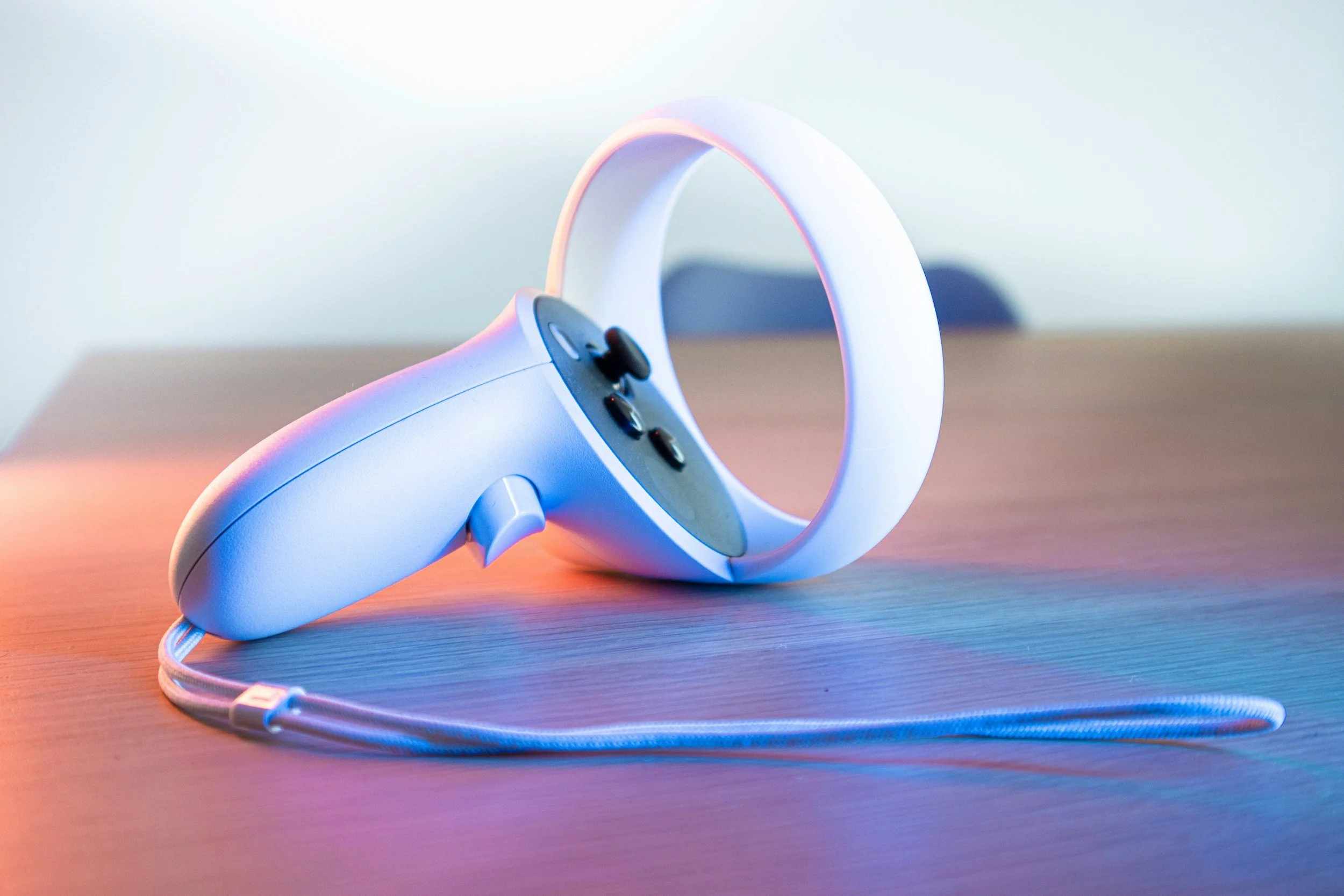 White VR headset with black and silver buttons lying on a wooden surface, cord attached, in front of a plain background with colorful lighting.