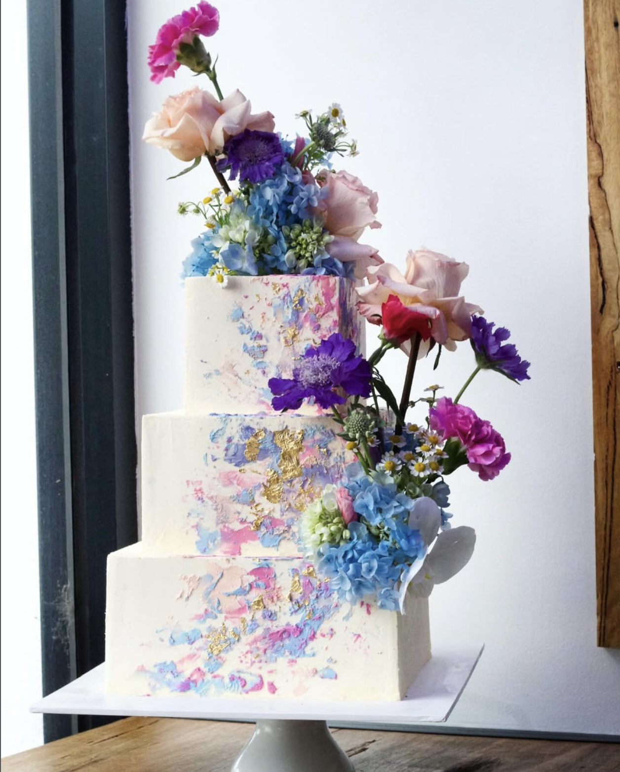 A decorated three-tiered cake with colorful floral accents and gold leaf details, placed on a white cake stand near a window.