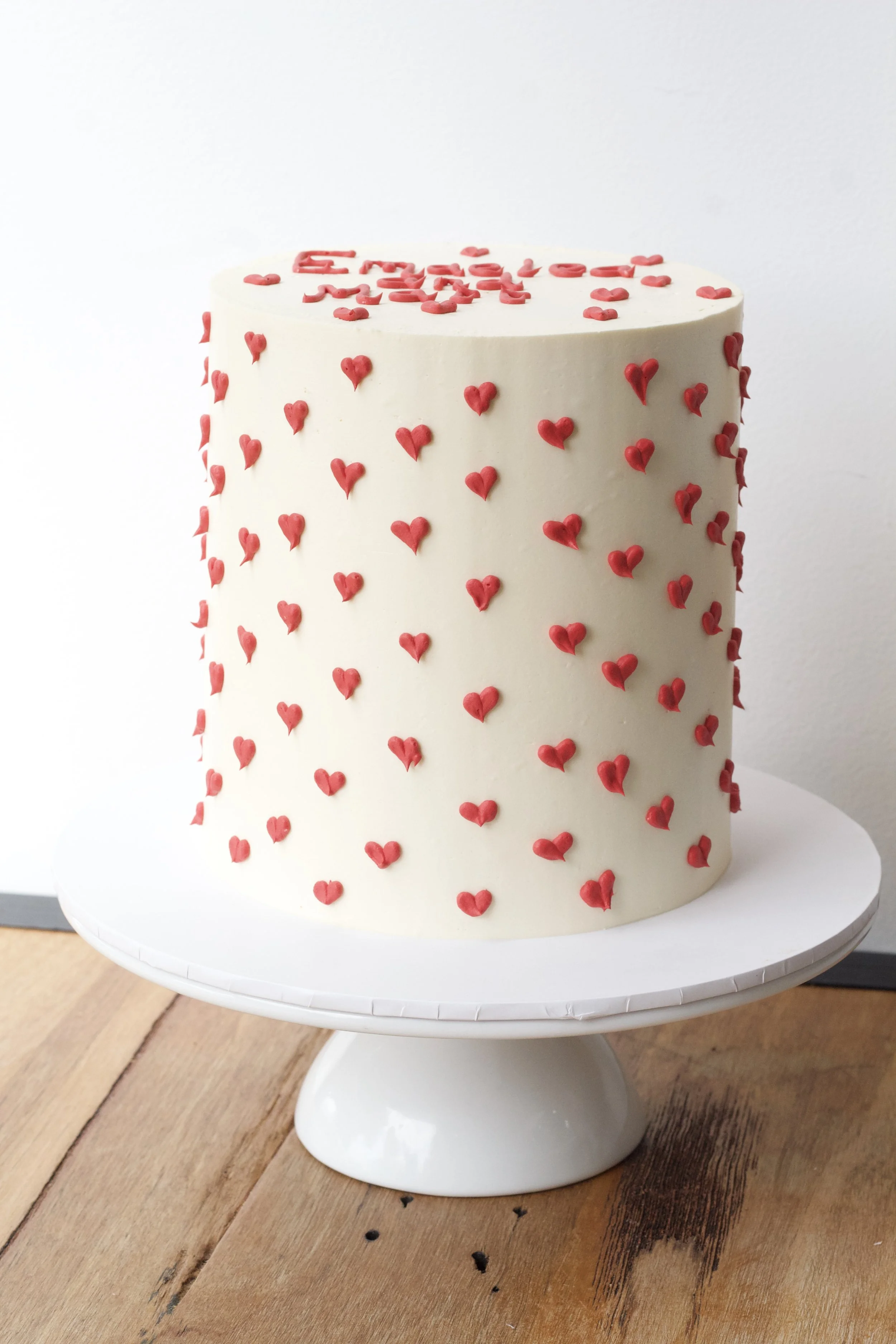 A white, heart-shaped cake decorated with small red hearts and some words written in red icing on the top, placed on a white cake stand on a wooden surface.