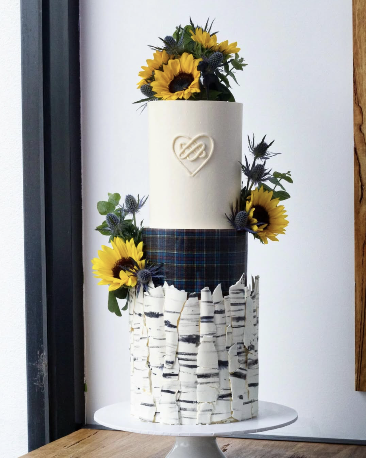 Multi-tiered cake decorated with sunflowers, blue thistle flowers, leaves, and a plaid ribbon. The cake features a white textured base, a middle tier with a tartan pattern, and an upper plain white tier with a heart and infinity symbol decoration.