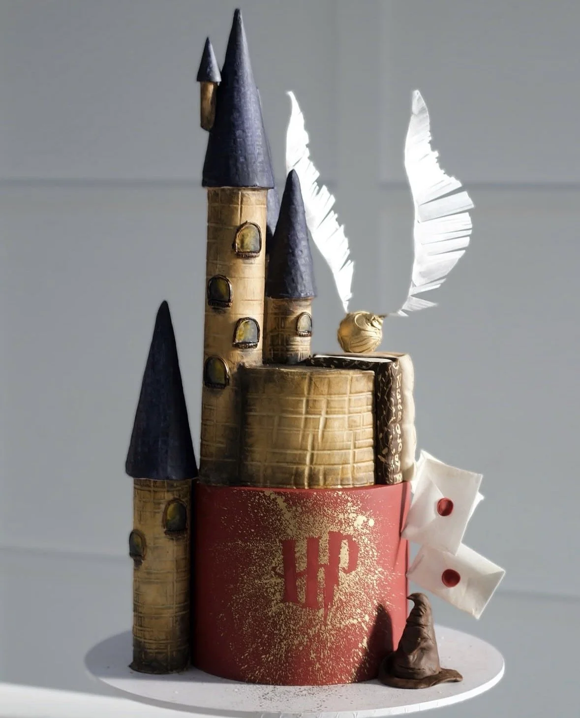 A cake designed to look like a castle, with a red base, towers with dark roofs, and a paper wing attached to the side, decorated for the harry P theme.