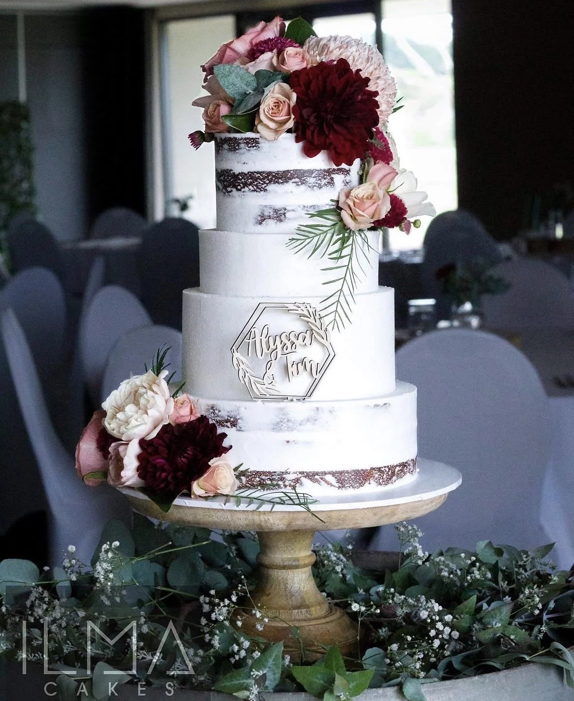 A three-tiered white wedding cake decorated with fresh pink roses, dark red dahlias, and greenery, placed on a wooden cake stand surrounded by flowers and greenery, with a wedding sign that reads "Alyssa and Tim."