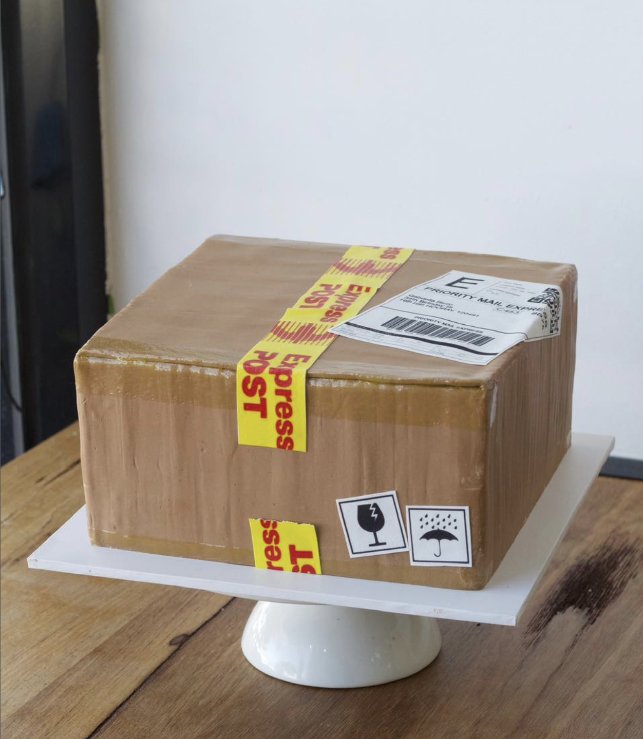 A cardboard box on a white cake stand, with 'Fragile' tape and shipping labels, including a USPS tracking label, and handling symbols for 'this side up' and 'keep dry'. 3D cake all edible and handmade with exceptional precision and attention details