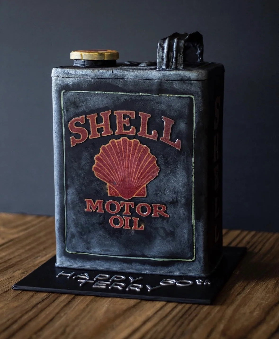 A cake shaped like a vintage Shell motor oil can, with detailed branding and design, sitting on a black base that says "Happy 40th Birthday" on a wooden surface.