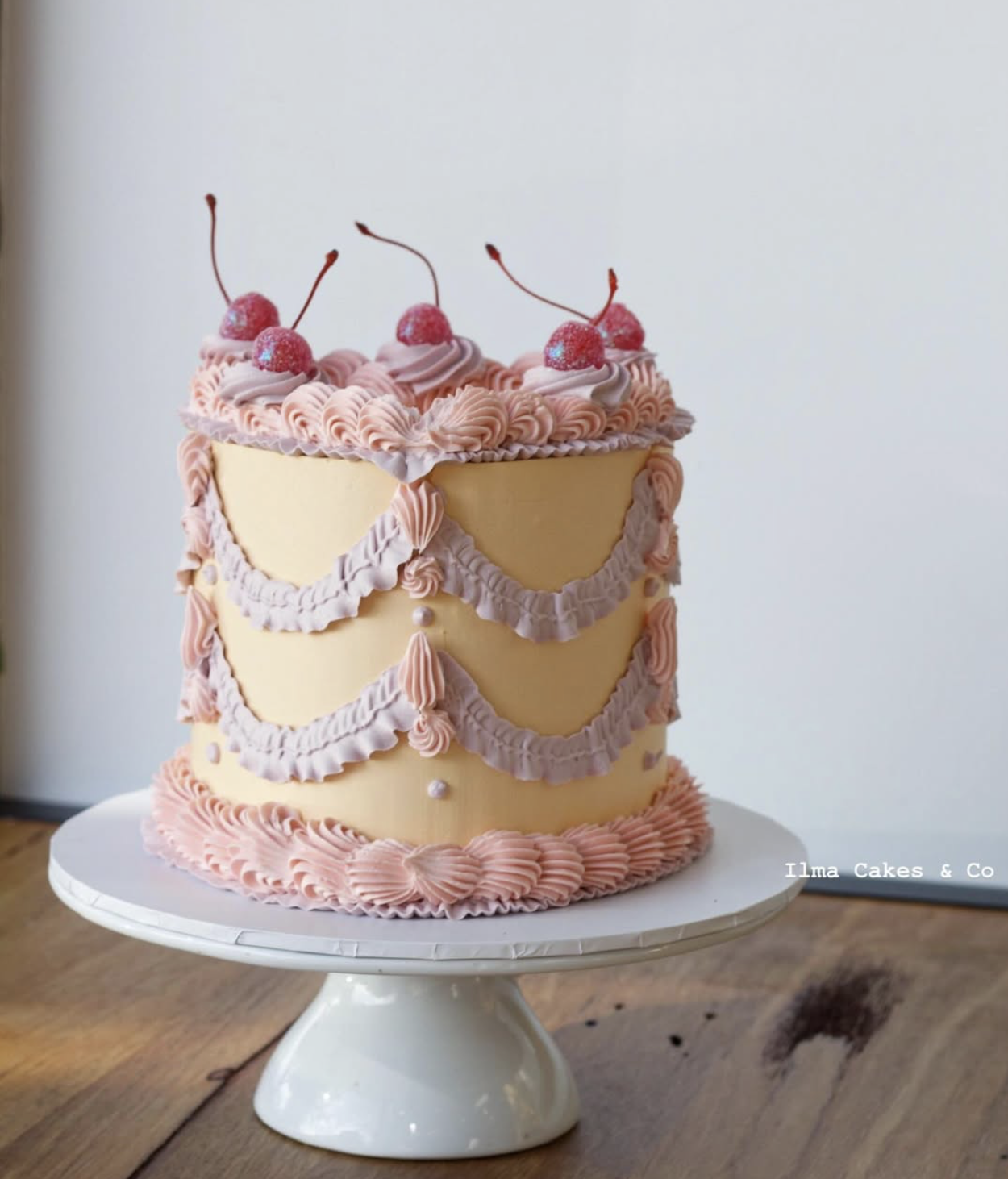 Decorative pink and yellow cake with cherry toppings, piped pastel-colored frosting, and pearl accents, placed on a white cake stand on a wooden table.