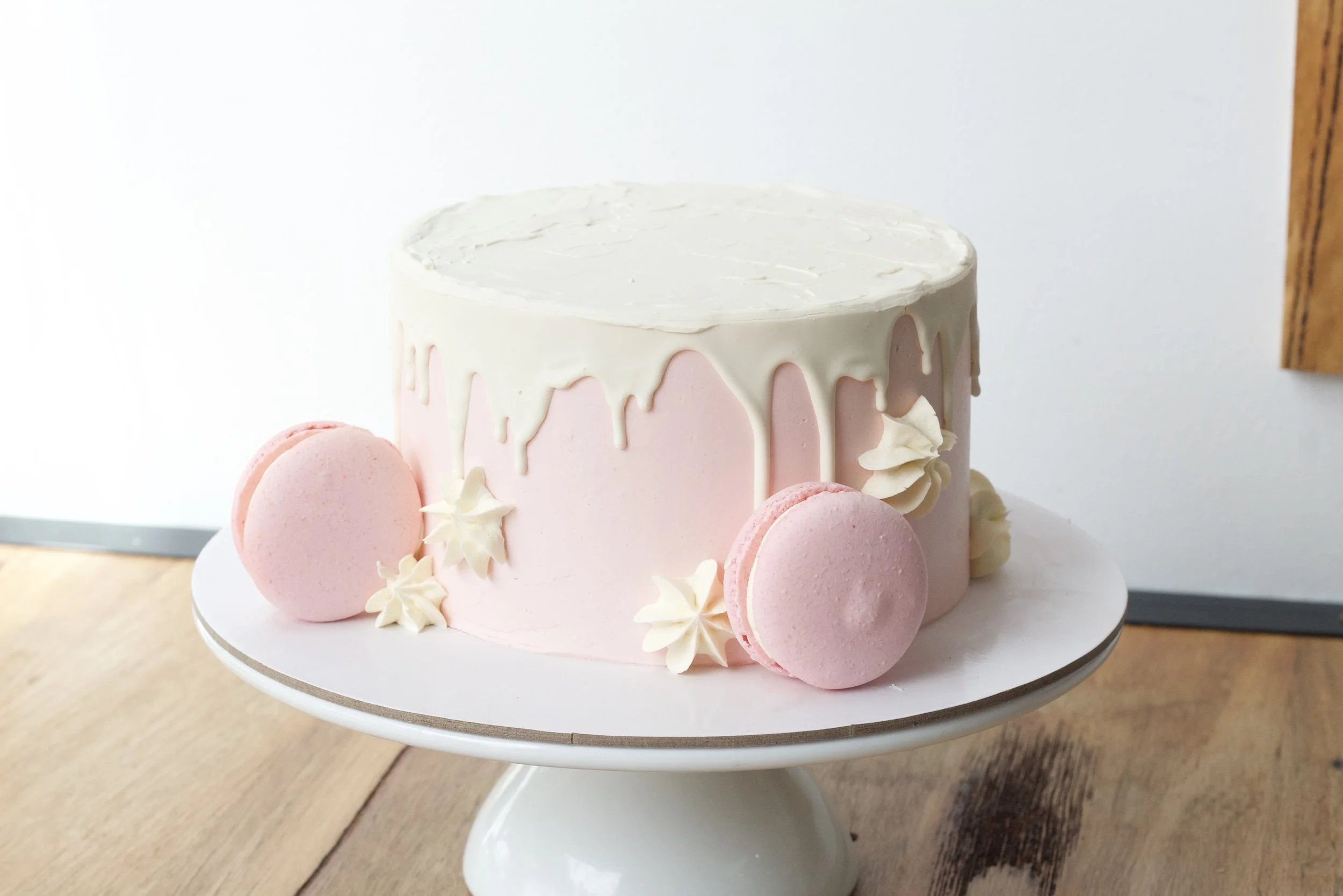 A pink and white cake with white icing dripping down the sides, decorated with pink macarons and white flowers, on a white cake stand.