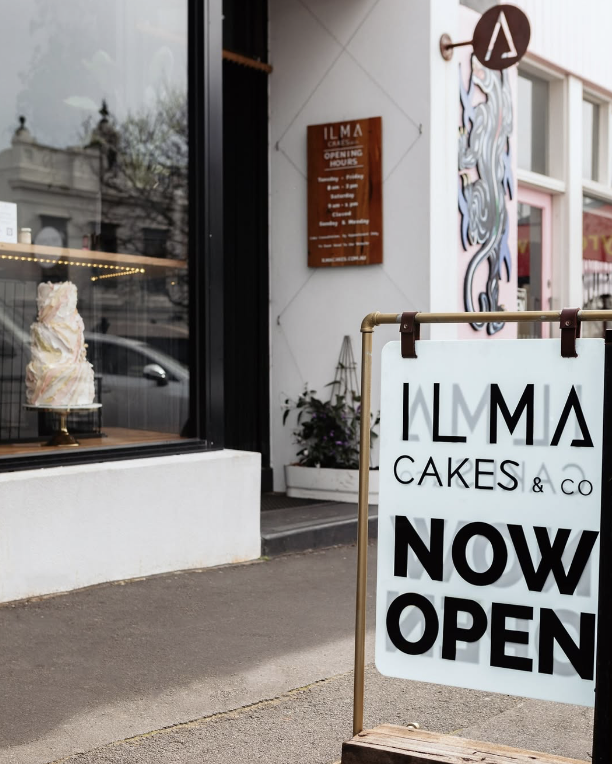 A front display of the Warrnambool based micro bakery with a white sign that says 'ILMA CAKES & CO NOW OPEN'. There is a sign on the sidewalk indicating the shop is now open, serving gourmet Toasties, pastries, focaccia and treatsl handmade on site