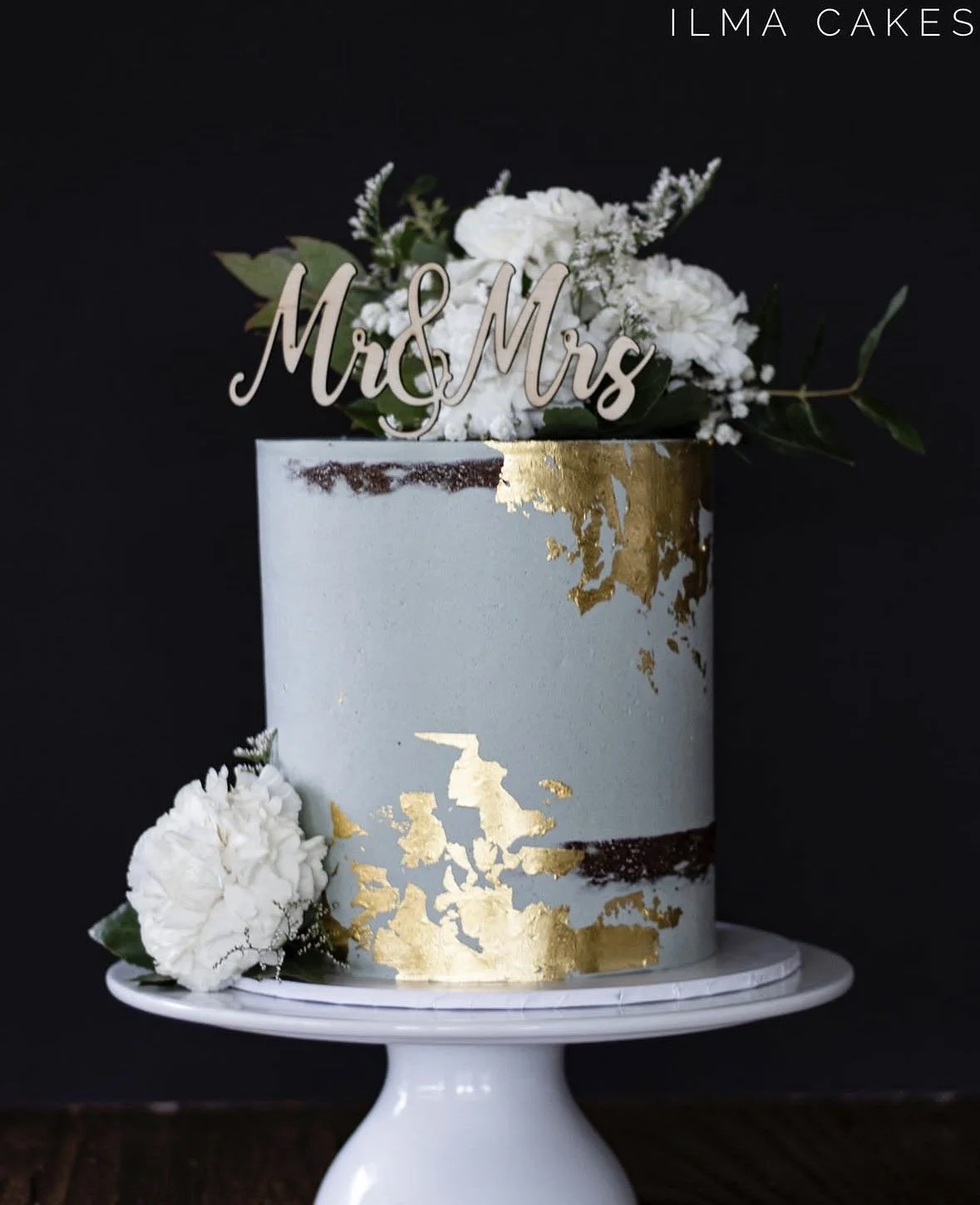 A elegant wedding cake with white and gold leaf accents, decorated with white flowers and a 'Mr & Mrs' topper.
