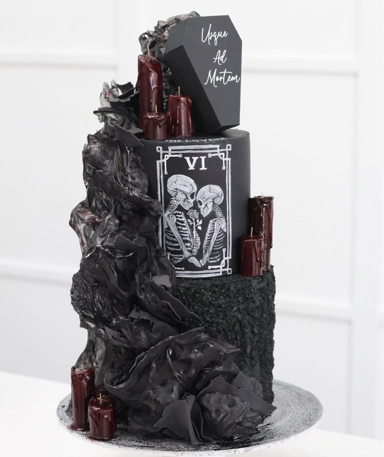 A gothic-themed cake with black, red, and dark gray colors, featuring skull and skeleton designs, topped with red candles and black elements resembling rocks or coal.