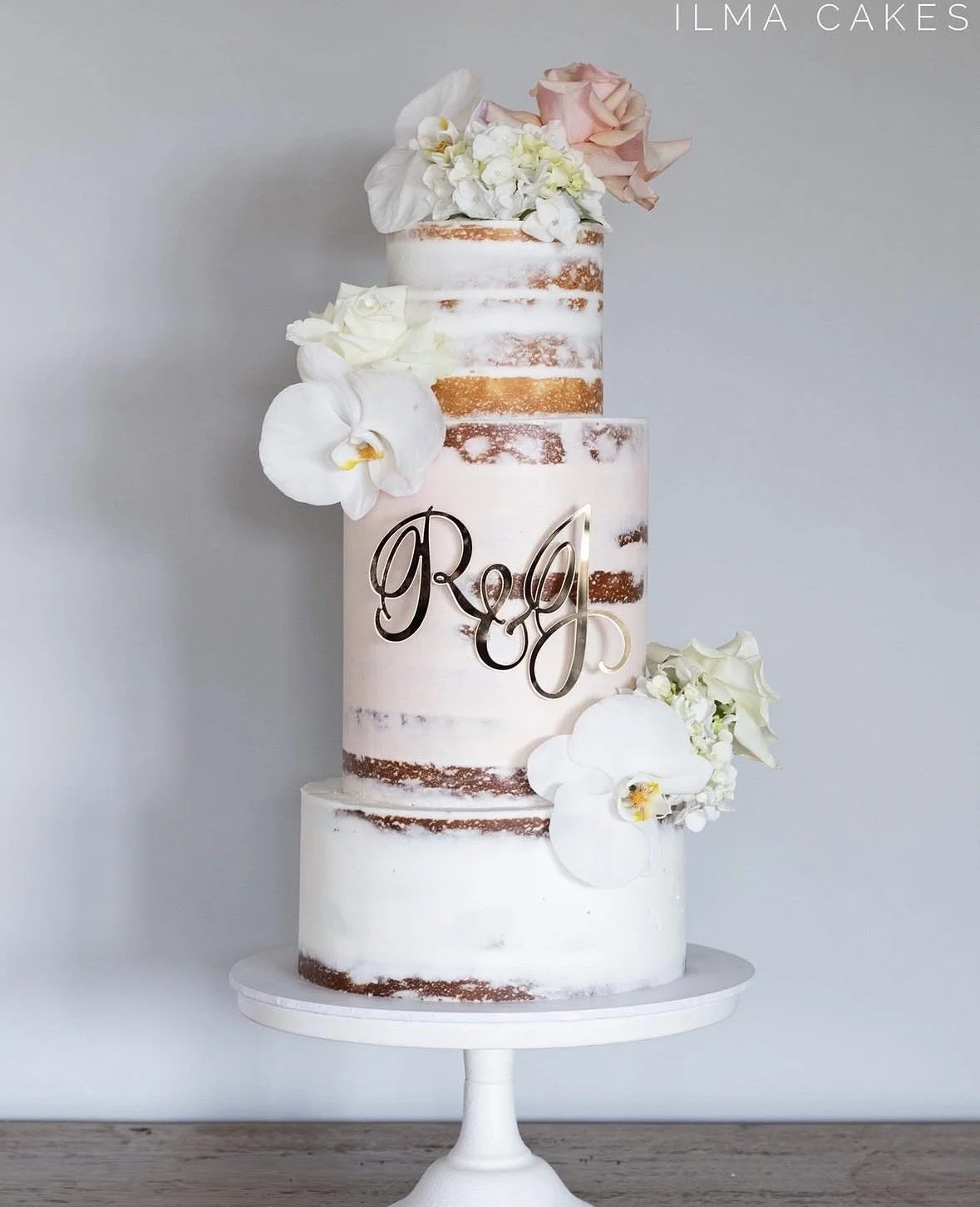 A three-tiered semi-naked wedding cake decorated with white and pink flowers and a gold monogram "REJ" on the front, placed on a white cake stand against a plain gray background.