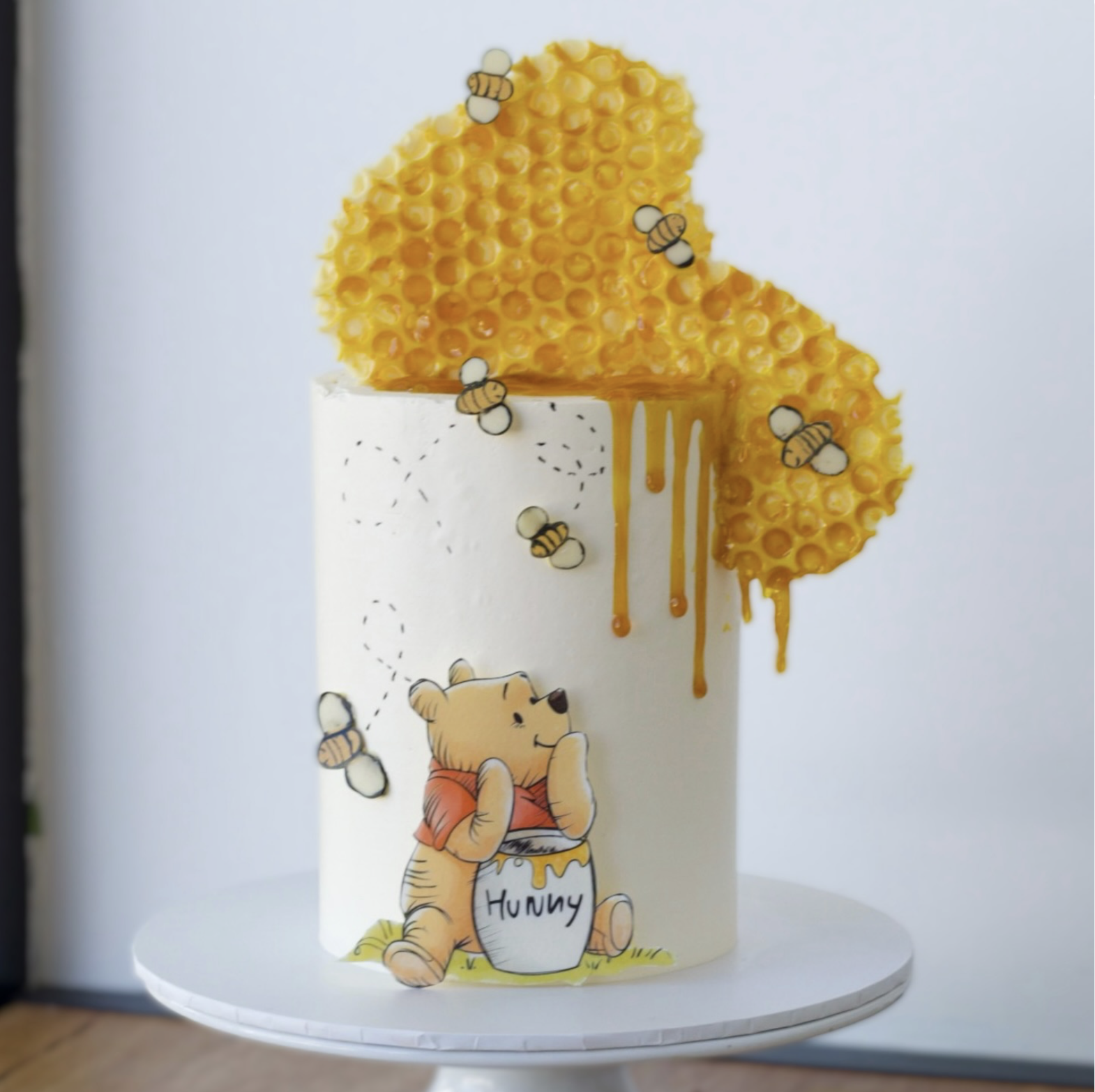 Cute illustration of Winnie the bear sitting on a white cake with honey jar, honeycomb, and bees around, evoking a playful and adorable theme.