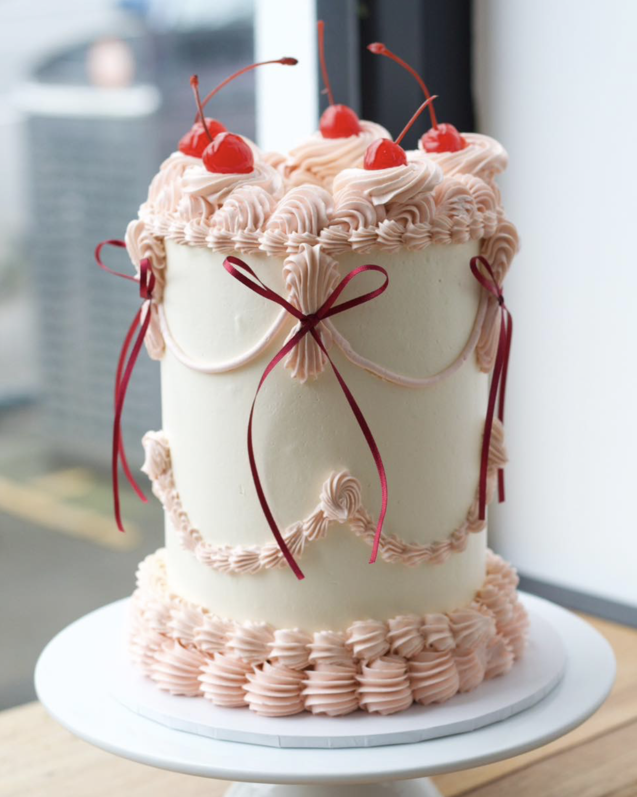 A tall, white birthday cake decorated with pink piped frosting, red cherries on top, and pink and red ribbons tied around the sides, sitting on a white cake stand near a window.