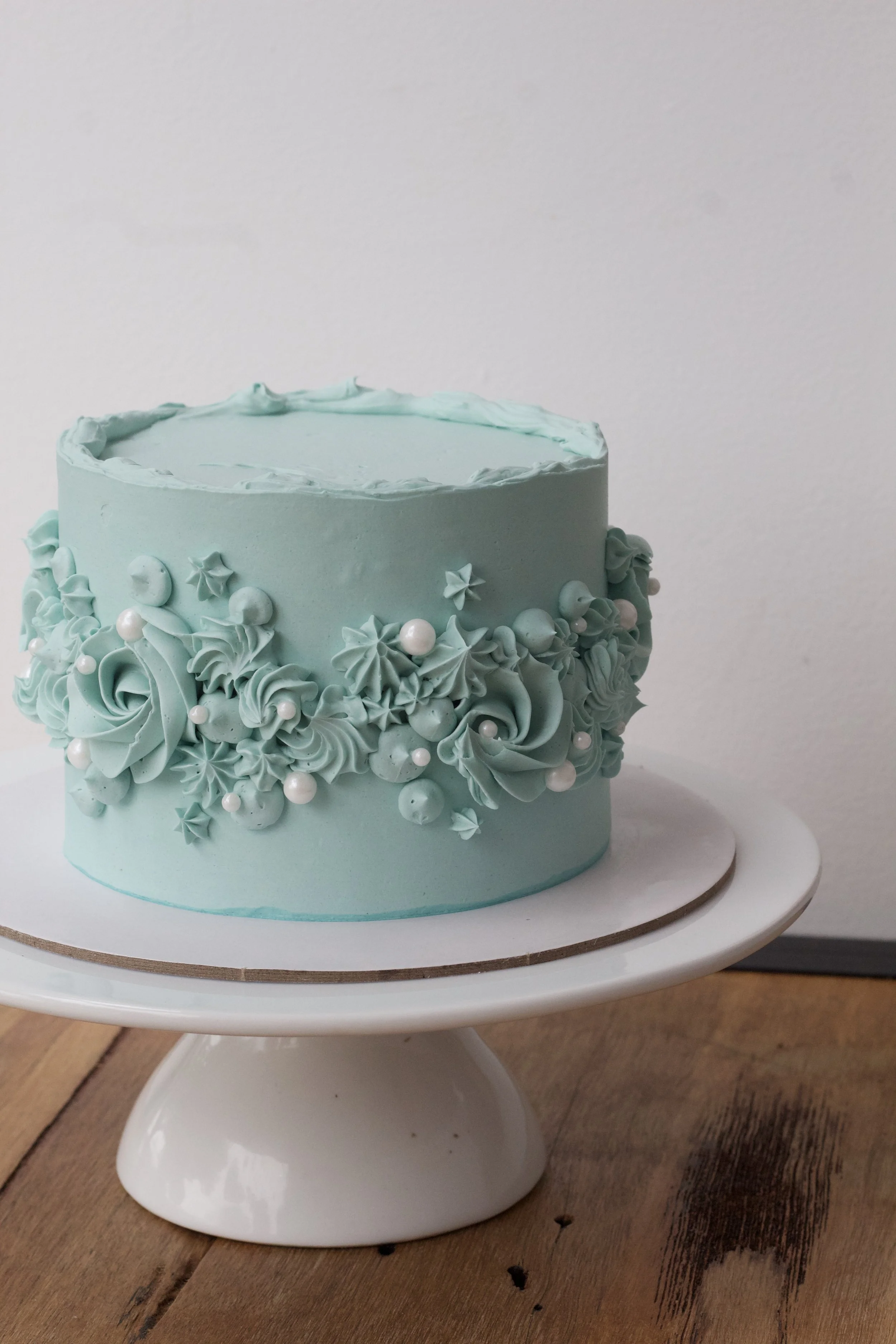 A round cake decorated with pale blue frosting, piped flowers, stars, and pearls, displayed on a white cake stand on a wooden surface.