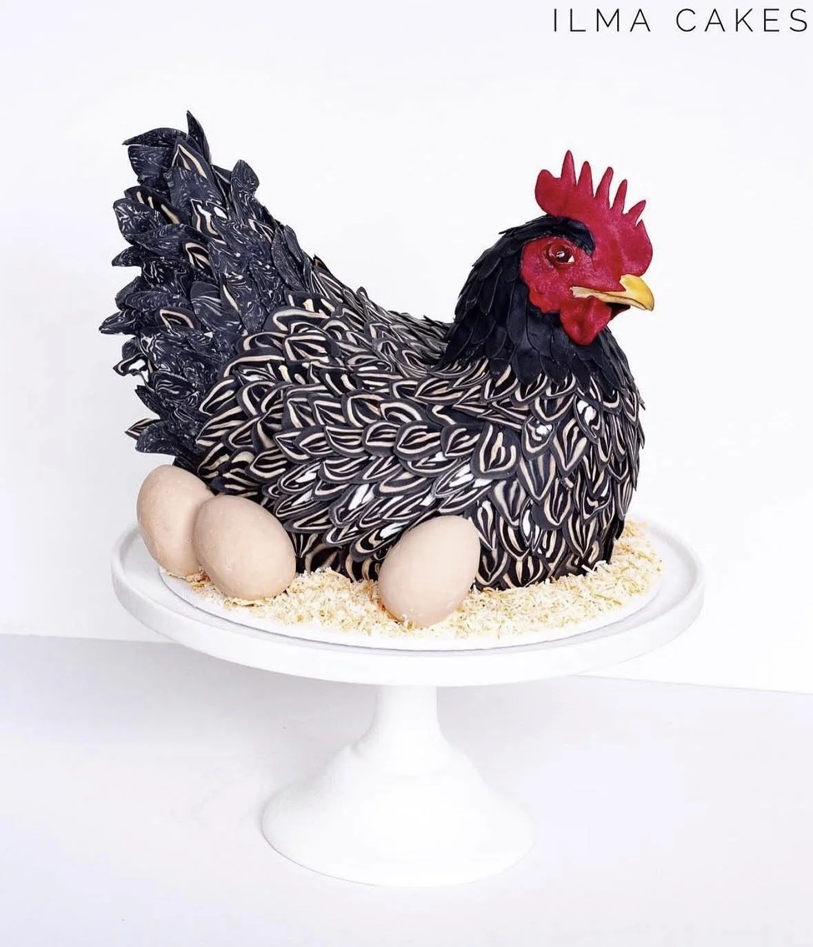 Cake shaped like a hen with black, white, and gray patterned feathers, red comb, and yellow beak, with three eggs at the base, on a white cake stand all handmade and edible as a gluten free mud cake but ultra realistic 