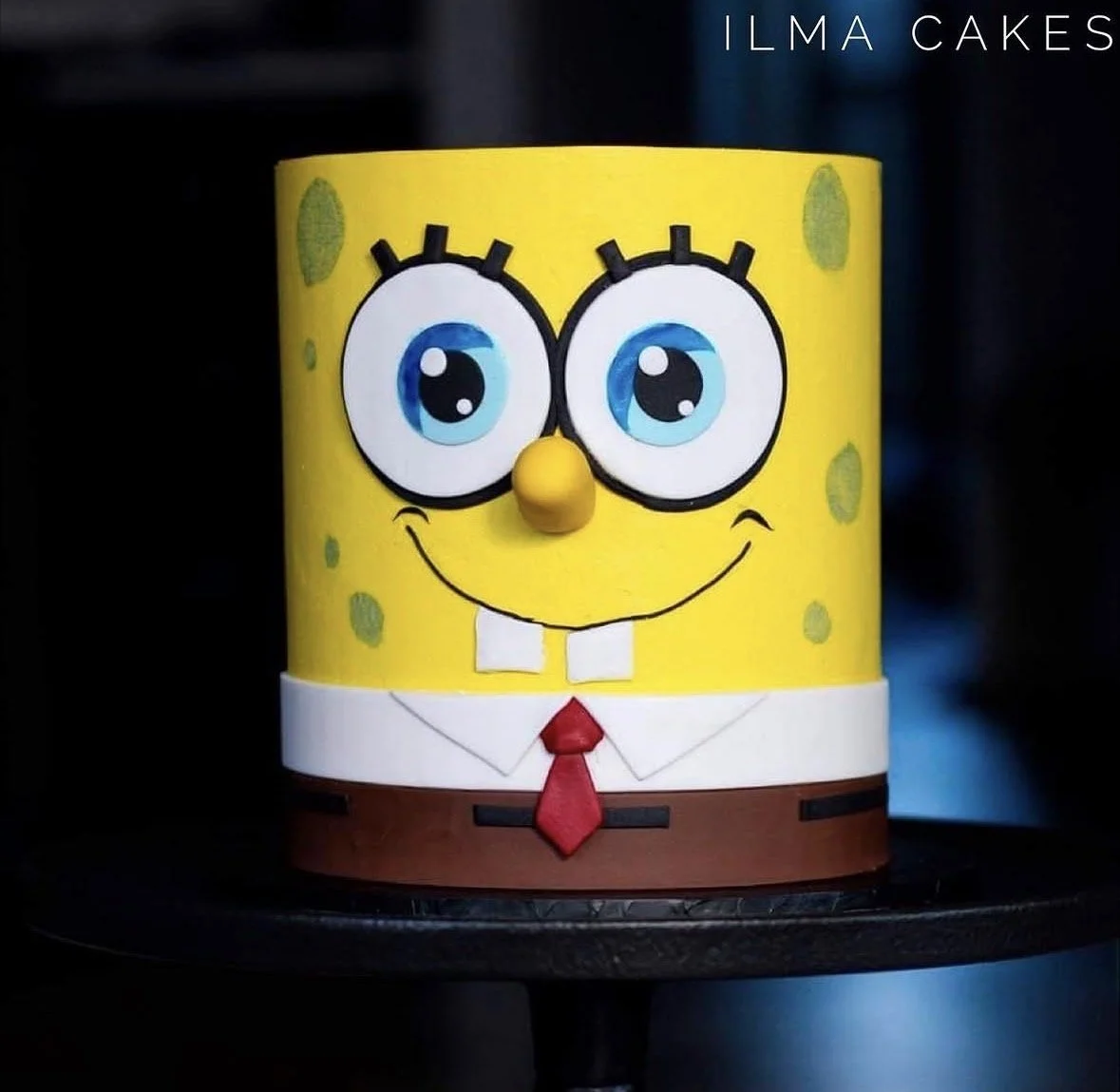 A cake designed to look like a cartoon character who is yellow with a sponge for a body  with yellow frosting, large blue eyes, a small yellow nose, and a red tie, placed on a black cake stand.