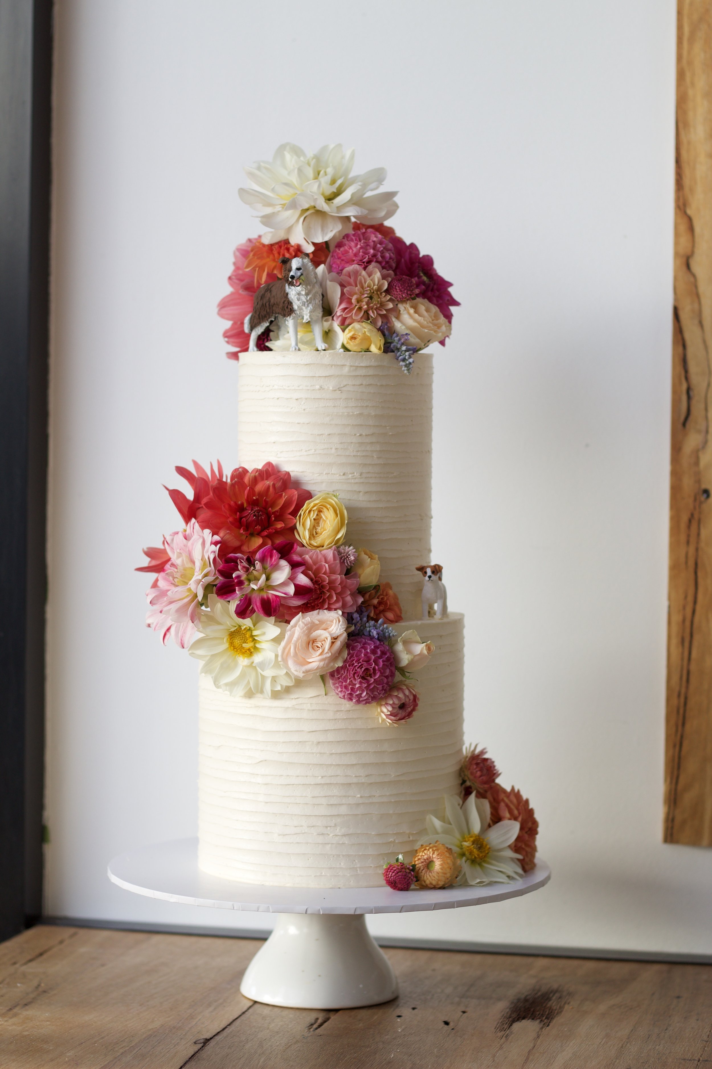 A tall white tiered cake decorated with a variety of colorful flowers and small dog figurines, placed on a white pedestal stand on a wooden surface.