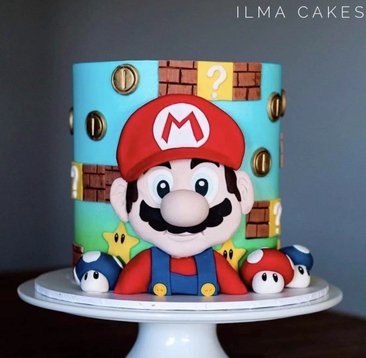 Decorative cake featuring a game theme with a large cartoon Mario figure in the front, colorful background resembling Mario's game environment, including coins, question mark blocks, and mushrooms, on a white cake stand.